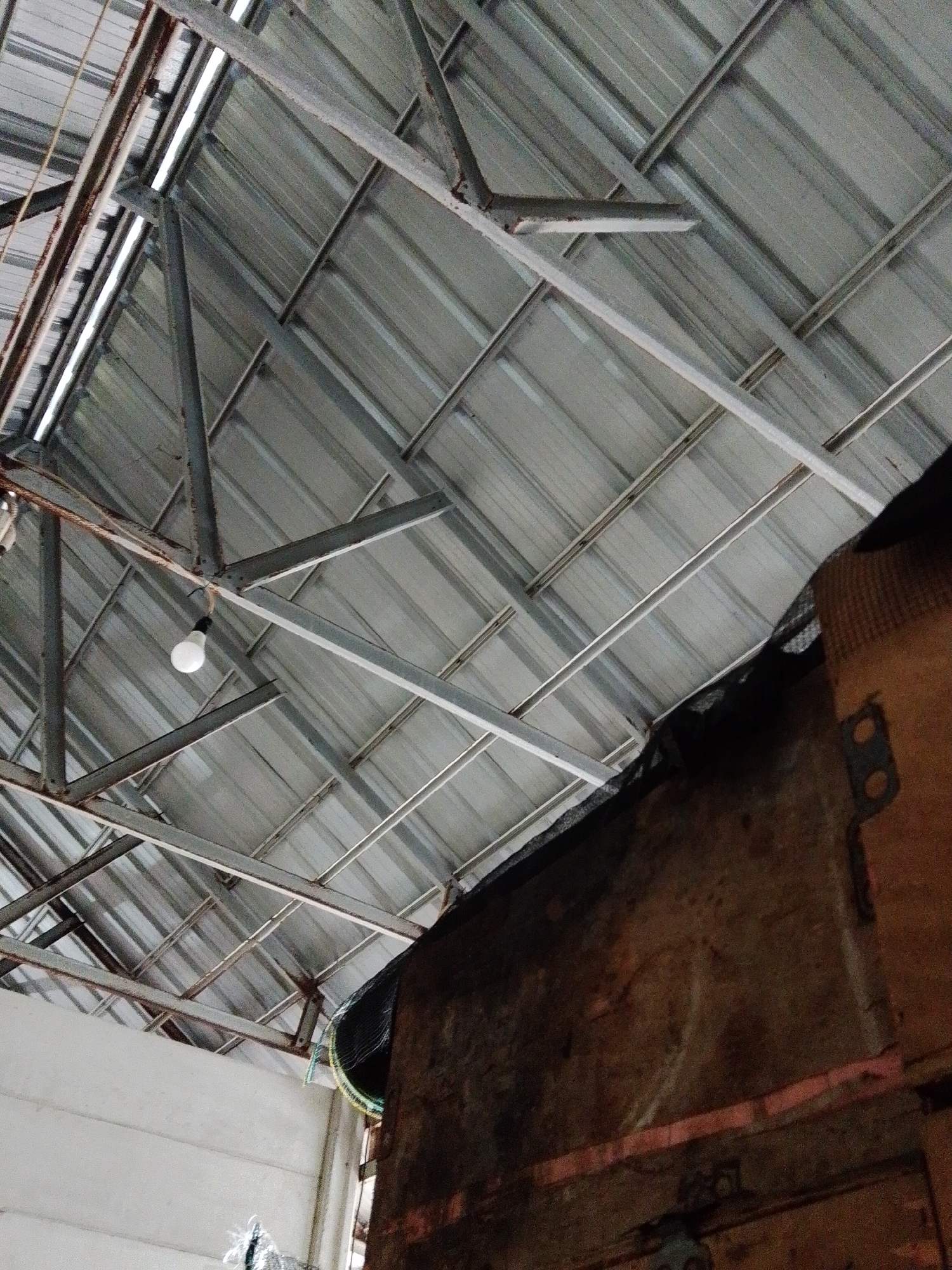 The image shows the interior of a building in Bukit Ketok, Indonesia. The roof consists of corrugated metal sheets supported by a metal framework. A single light bulb hangs from the framework. A wooden wall is visible on the right side of the image, with netting attached near the ceiling. The overall environment appears to be an industrial or storage space.