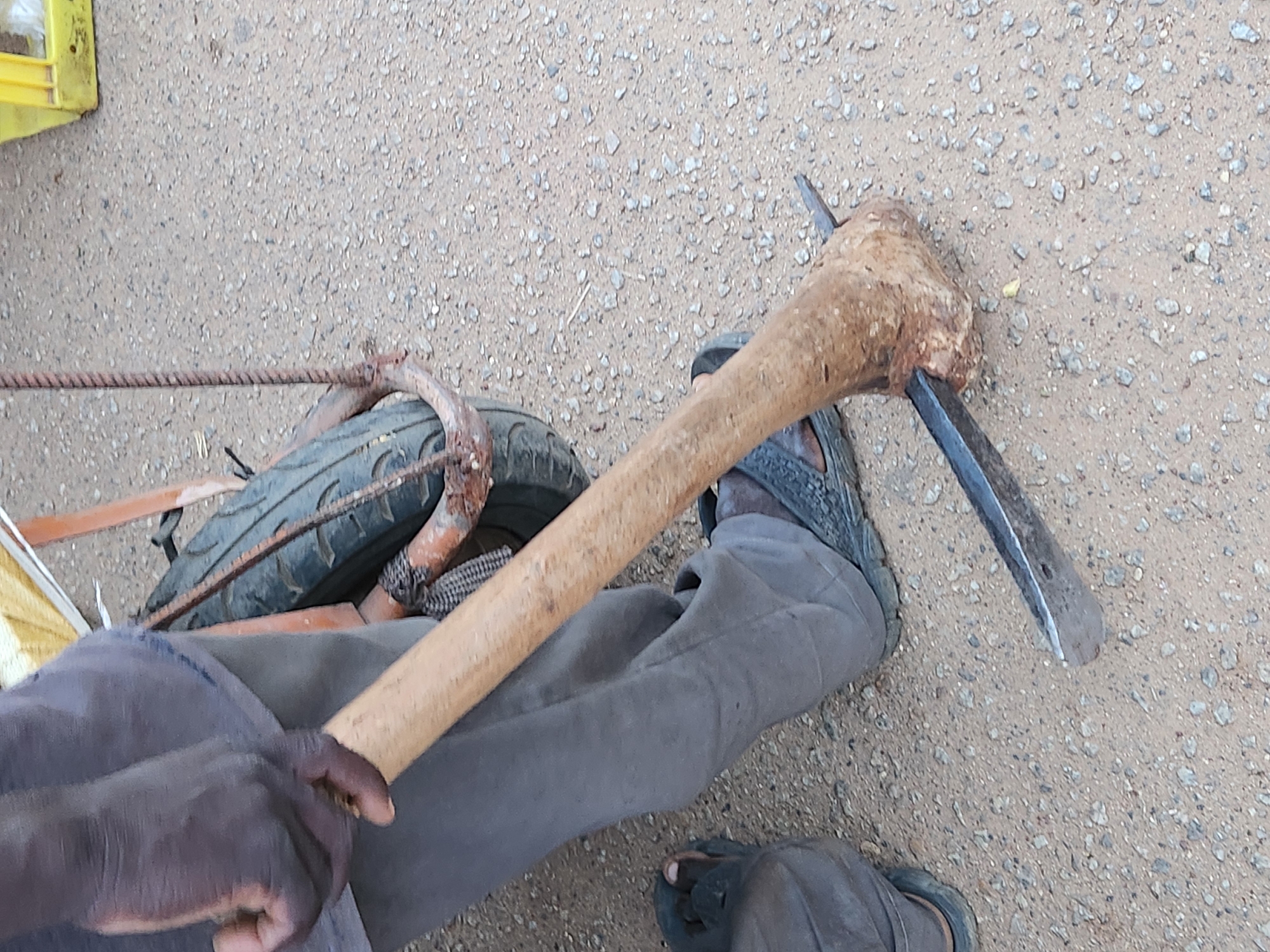 In Kabong, Nigeria, a person wearing grey trousers and dark sandals is holding a pickaxe. The pickaxe has a wooden handle and a metal head. The person's left hand is gripping the handle near the head. The scene appears to be outdoors on a gravelly surface, with a tire and part of a cart visible in the background.
