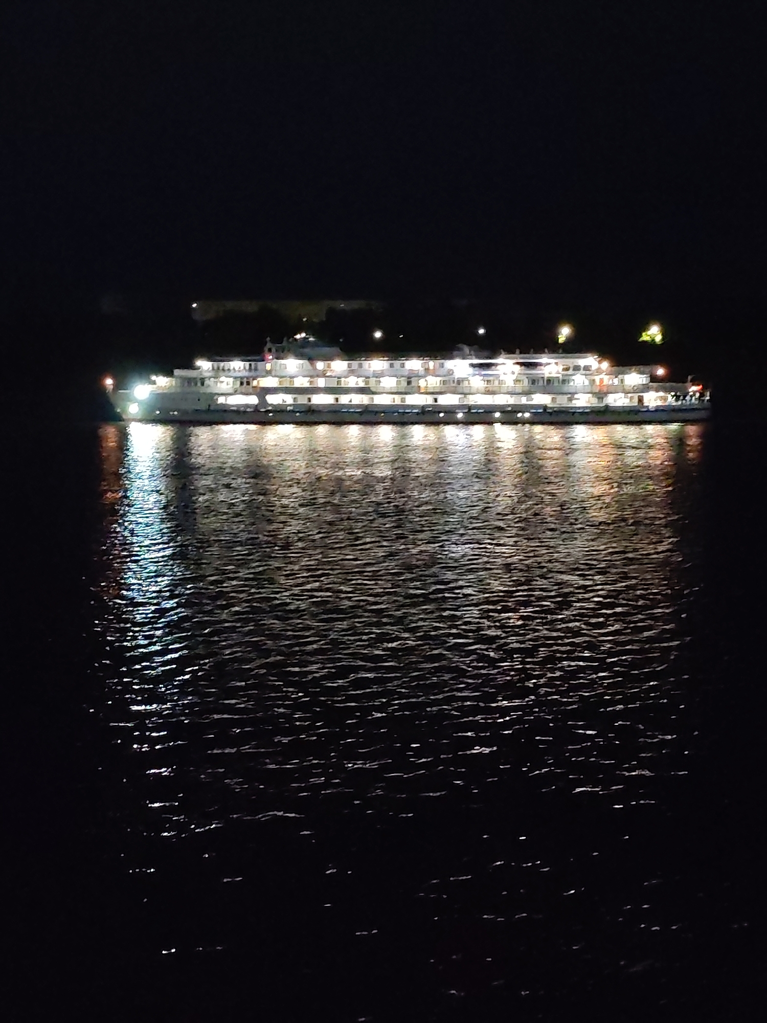 The image shows a large white cruise ship illuminated with lights at night, docked on a body of water. The ship is long and multi-decked, with numerous windows and lights visible on its sides and top levels. The water reflects the ship's lights, creating a shimmering effect. The background is dark, indicating nighttime, with a faint silhouette of land and trees on the horizon, possibly indicating a riverbank or shoreline. The scene is likely a port or mooring area.