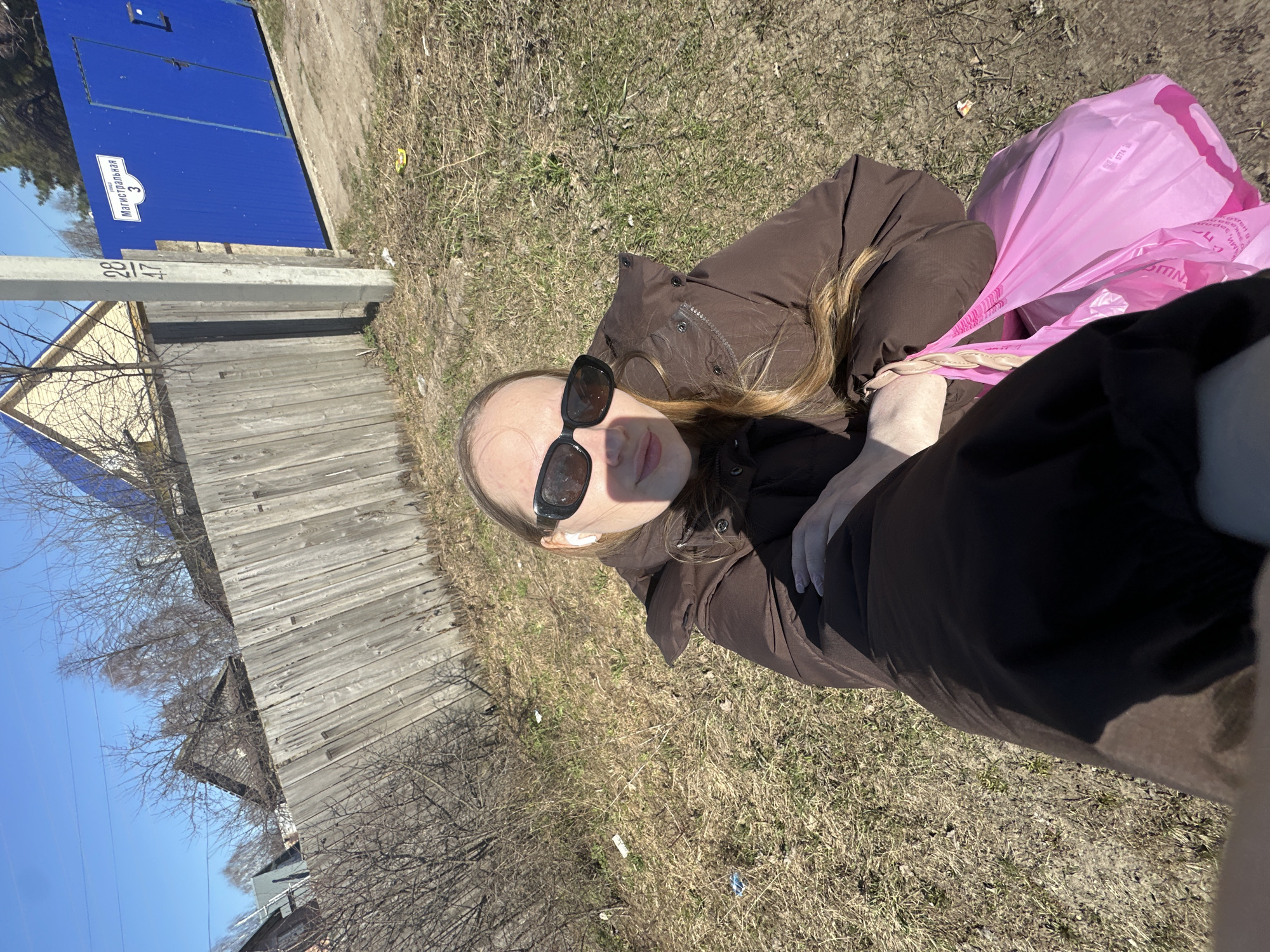 The image shows a woman wearing sunglasses and a brown jacket, holding a pink bag. She is lying on the ground with dry grass. Behind her is a gray wooden fence, a blue gate with a street sign, and a utility pole with numbers. Trees and a blue sky are visible in the background. The street sign reads "Магистральная 3" (Magistralnaya 3). Based on this street name, the location is in the city of Chaikovsky, Perm Krai, Russia.
