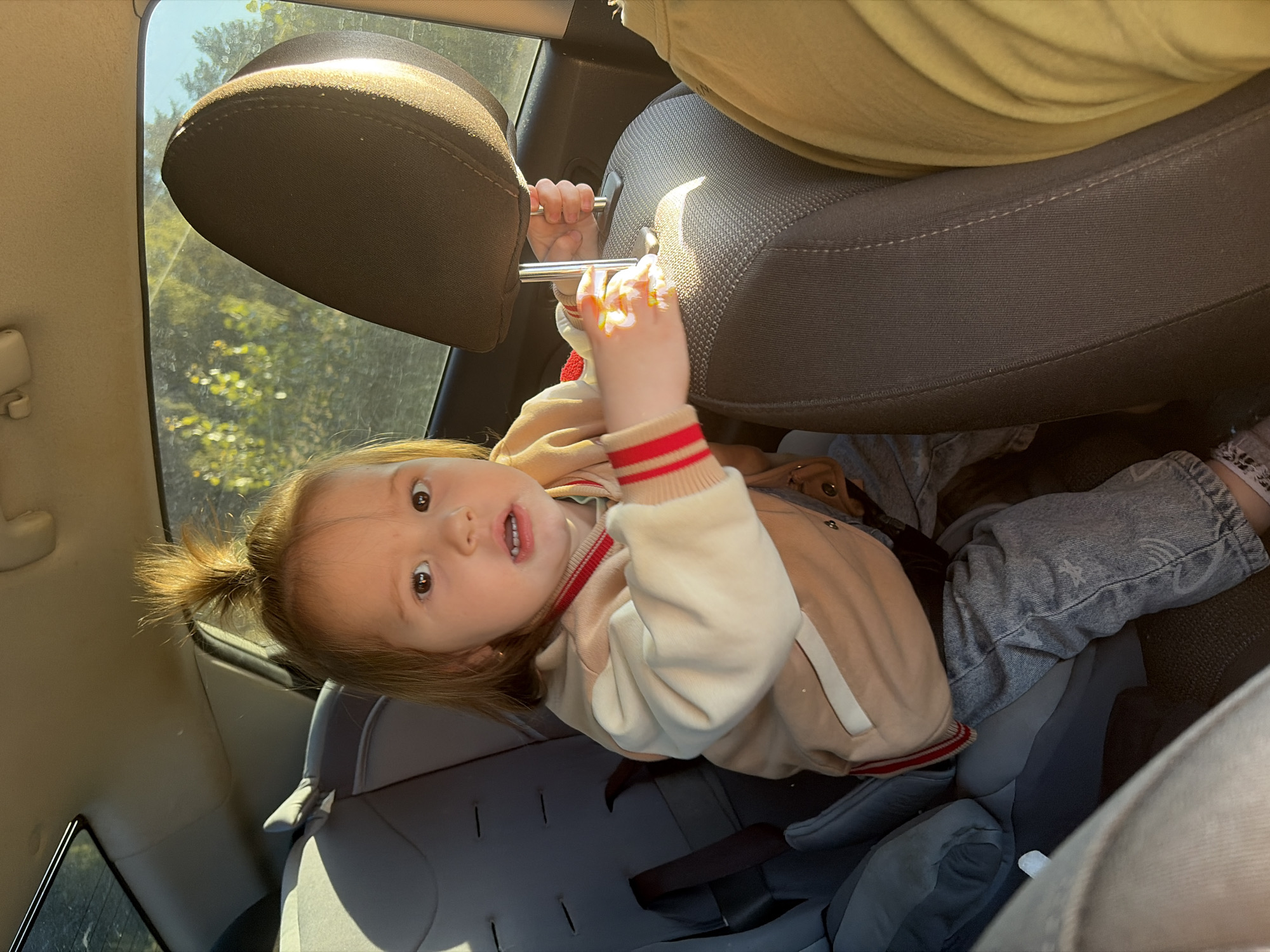 A young child with light brown hair tied in a bun is sitting in a car seat in the back of a car. The child is wearing a light tan jacket with red and white striped cuffs and collar, and light blue jeans. The child is looking towards the camera with a slightly surprised expression, their mouth open. The child's hands are resting on the headrest of the front seat. Sunlight streams in from the side window, illuminating the scene. Outside the window, trees and foliage are visible, suggesting a rural or suburban setting. The interior of the car appears to be a standard vehicle. The specific location of Chernushka, Russia, is not discernible from the visual information in the image.