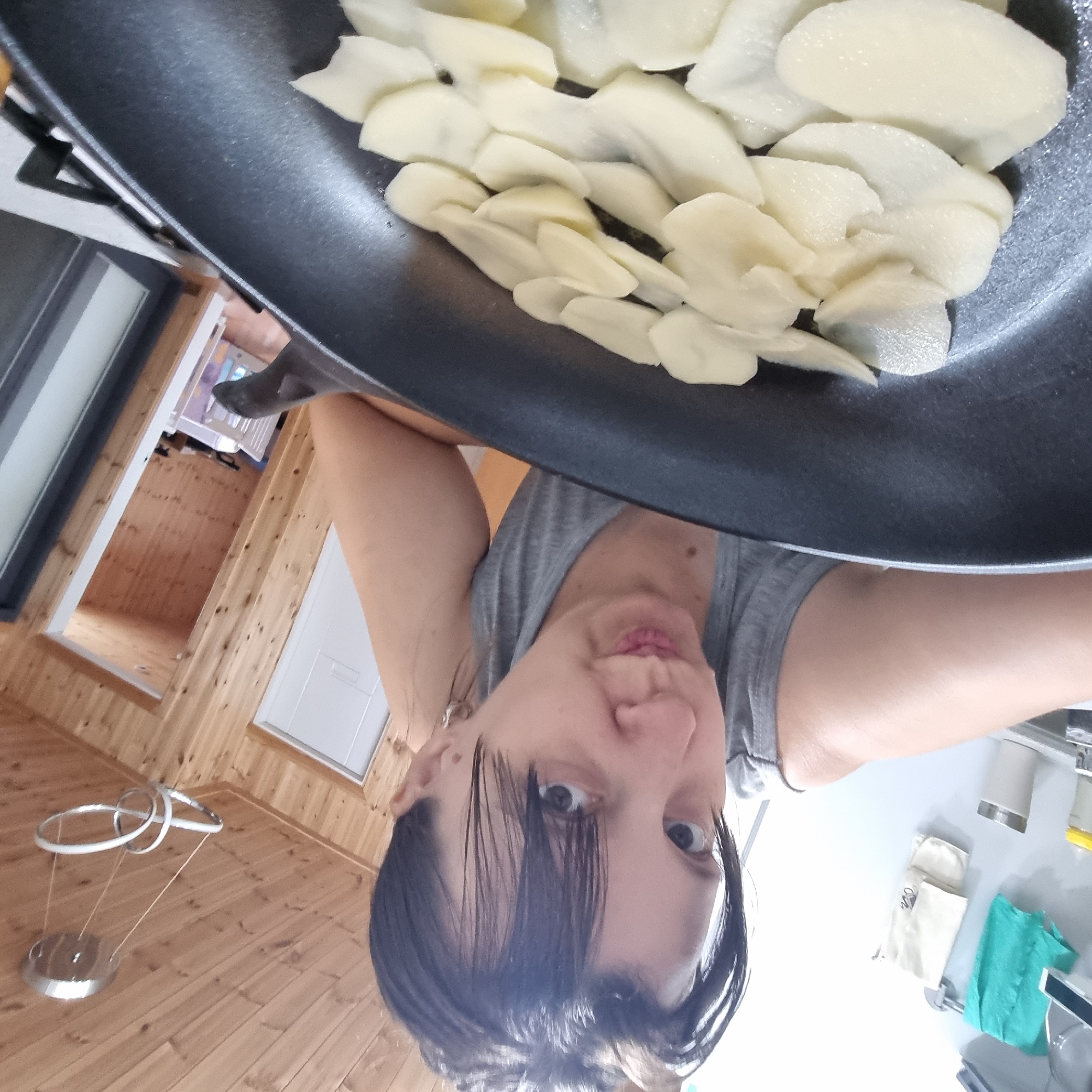 An over-the-shoulder, selfie-style shot captures a woman looking at the camera while leaning over a black frying pan filled with thinly sliced potatoes. The woman has dark hair, bangs, and is wearing a grey tank top. She appears to be in a kitchen with wooden paneled walls and ceiling. A modern light fixture hangs from the ceiling. In the background, a doorway leads to another room with wooden walls and a small white baby crib. The scene is illuminated by indoor lighting.