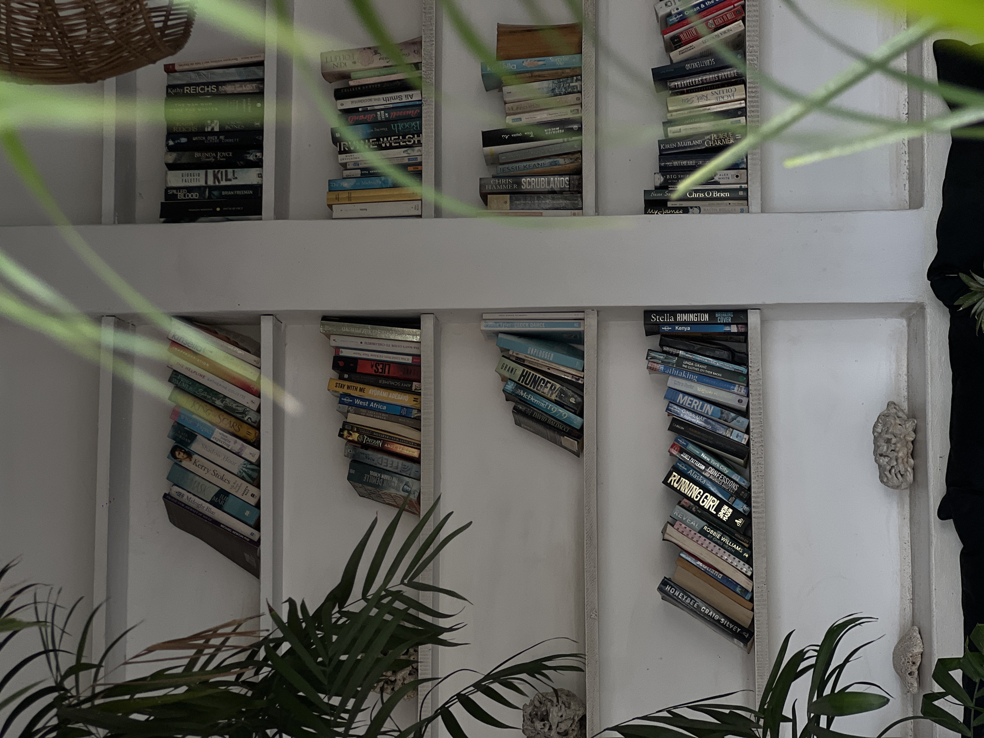 The photograph depicts a white bookshelf with multiple cubbies filled with stacked books. The books are oriented vertically and arranged haphazardly, with titles facing forward. The bookshelves are partially obscured by lush green foliage, suggesting an indoor setting with plants. The background is white and appears to be a wall. Some decorative items, such as a woven basket and carved stones, are also visible. The picture was taken indoors, possibly in North Kuta, Indonesia, as indicated in the prompt. No people are visible in the frame. The focus is on the bookshelves and the interplay of books and greenery.
