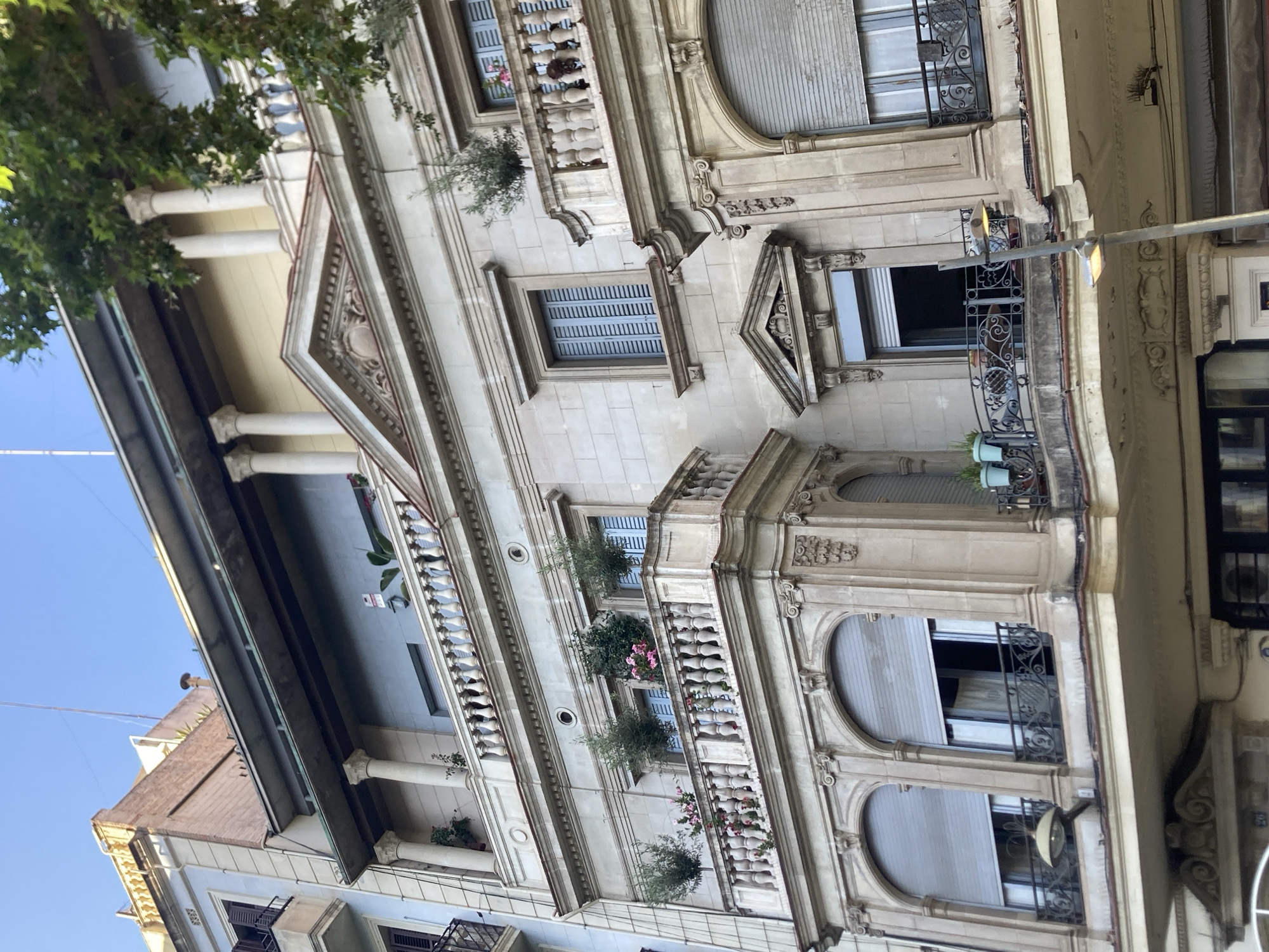 This is a low-angle, eye-level, outdoor, daytime, medium shot of the facade of a multi-story building in Barcelona, Spain. The building appears to be old, with ornate architectural details. The facade is primarily light-colored stone, with decorative columns and balconies. Several windows are visible, some with closed shutters and others open, revealing glimpses of curtains or blinds. Small potted plants are placed on the balconies. A streetlamp is positioned in the foreground, extending diagonally across the frame. The sky is visible at the top of the image, with a clear blue hue. A tree branch with green leaves extends into the upper right corner of the frame.