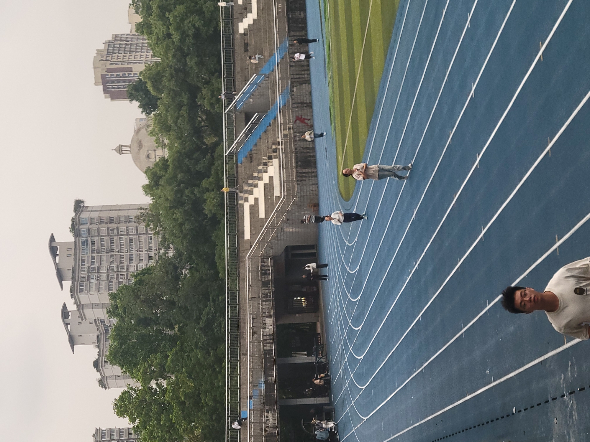 The image depicts a running track in the Shapingba District of China. The track is blue with white lane markings, adjacent to a green field. Multiple people are present on the track and in the adjacent bleachers. Several buildings are visible in the background, partly obscured by trees. The sky is overcast.