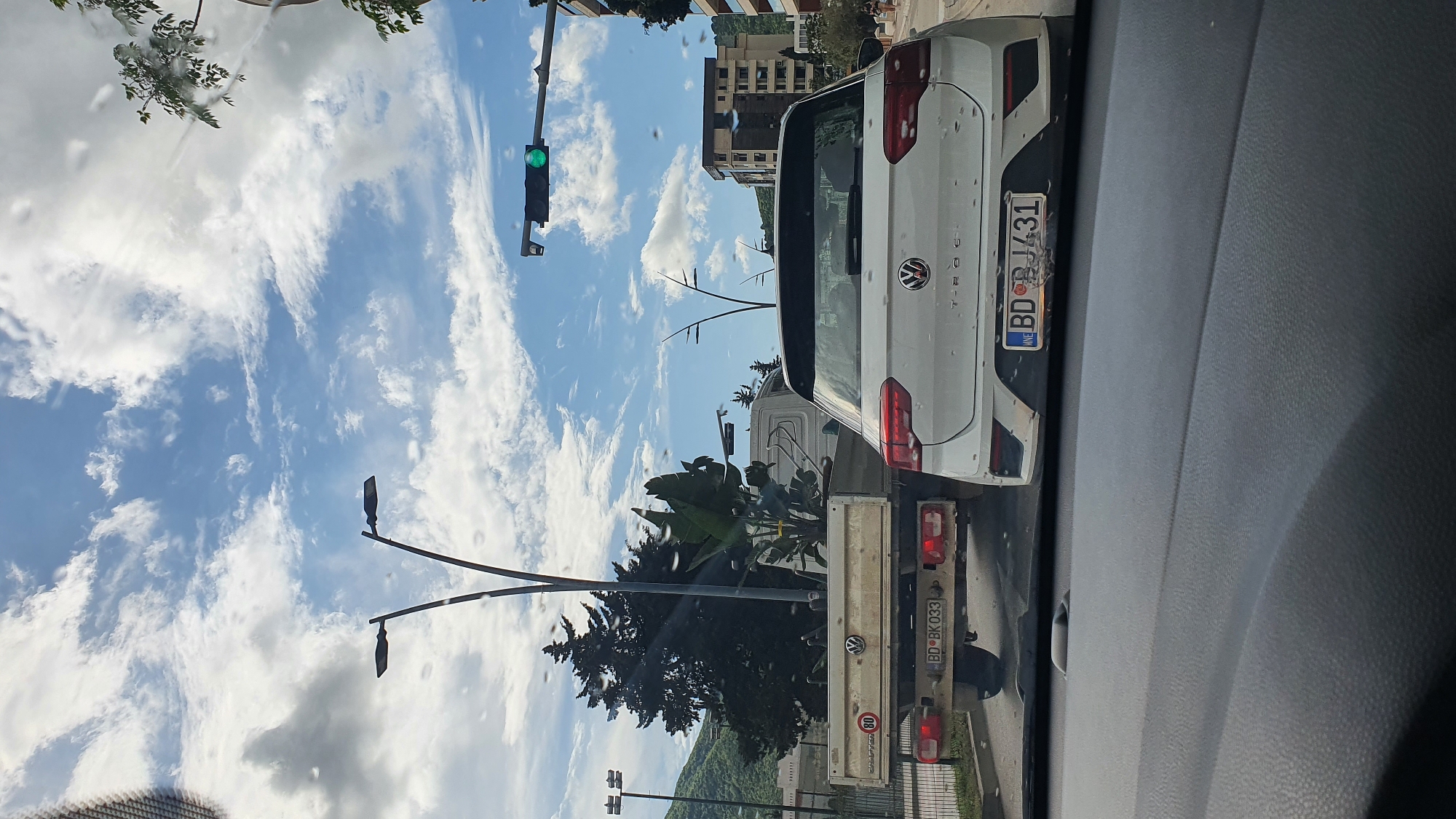 The image presents a daytime street scene from the perspective of someone inside a vehicle, looking through a windshield speckled with what appear to be water droplets or smudges. The sky is a prominent feature, displaying a bright blue expanse filled with dramatic, elongated white clouds.
In the foreground, a white Volkswagen T-Roc SUV is seen from the rear. The "VOLKSWAGEN" and "T-ROC" emblems are clearly visible on its tailgate. Its Montenegrin license plate reads "BD BD J431", with the country code "MNE" on the blue strip. Immediately behind the T-Roc is a white Volkswagen flatbed truck, also seen from the rear. Its license plate is "BD BK 033", and it has an "MNE" country code. A circular speed limit sticker showing "80" is affixed to the truck's rear. The truck's flatbed appears to be carrying large green plants.
To the left of the cars, a traffic light is clearly visible, illuminated green. Several street light poles with modern fixtures are scattered throughout the frame. Green trees with lush foliage and some with bare branches line the street. In the middle ground, buildings with multiple stories are visible, suggesting an urban or suburban setting. Distant mountains rise in the background under the cloudy sky.
The scene depicts traffic stopped, likely at an intersection, with the green light indicating permission to move. The bright sunlight and clear sky suggest it's a sunny day. The "BD" prefix on the license plates confirms the location as Budva, Montenegro.