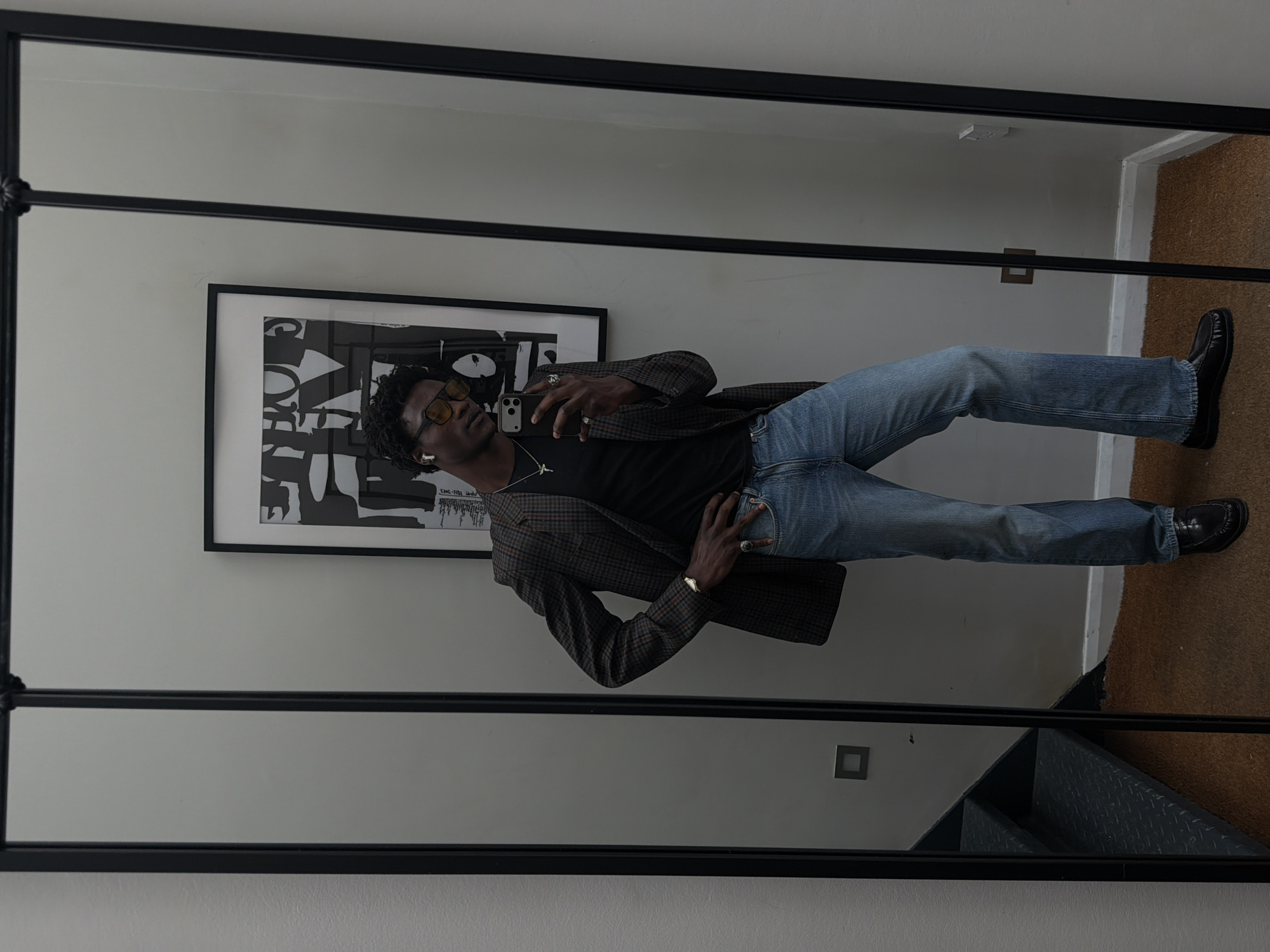 A medium, indoor shot depicts a reflection of a slender, dark-skinned male, likely in his late 20s to mid-30s, taking a selfie in a large, rectangular, black-framed mirror. The man is dressed in a casual yet stylish manner, wearing a black t-shirt, a patterned blazer in tones of brown and grey, light blue jeans, and black shoes. He also sports translucent yellow sunglasses, a gold chain necklace, and a watch on his left wrist.
The reflection shows the man holding his smartphone in his right hand, presumably to capture the image. He is positioned against a white wall, upon which hangs a framed piece of abstract black and white art. An electrical outlet is visible above and to the right of the artwork.
The lower right corner of the mirror's reflection reveals a section of stairs with a metal railing and a carpeted landing. The entire scene gives the impression of a modern, minimalist interior, possibly within a residential building. While the image provides no direct indications of location, the aesthetic and architectural elements are suggestive of an urban setting, such as London, United Kingdom. However, pinpointing a specific location within London is not possible based solely on the provided image.