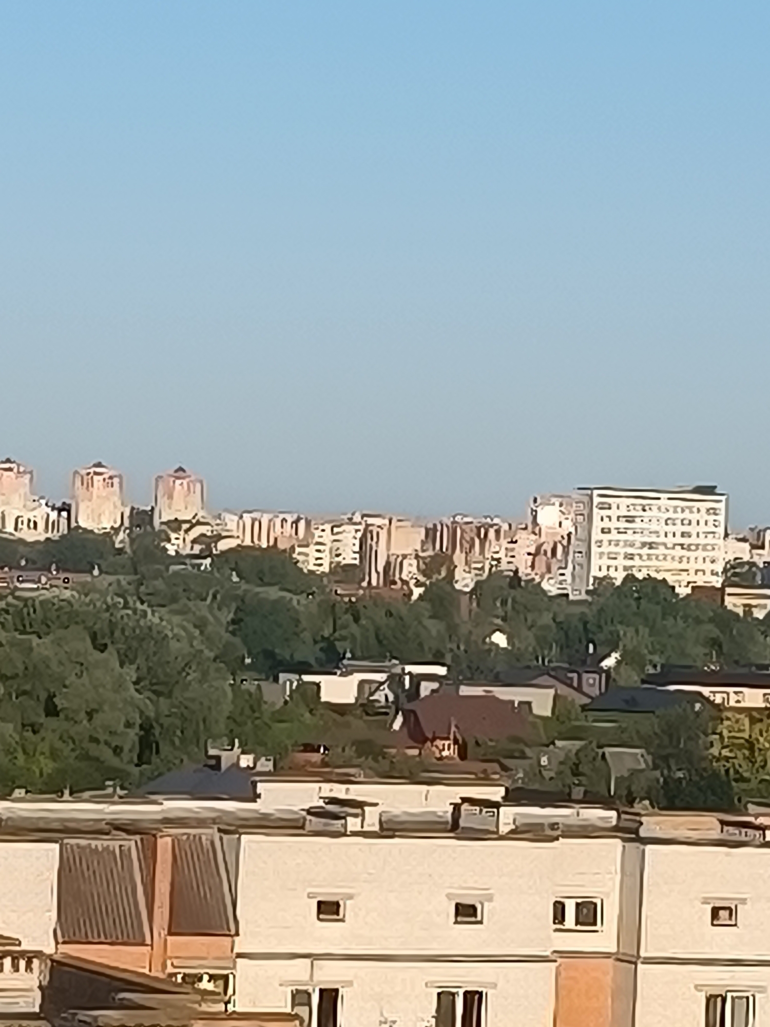The image shows an urban skyline with several multi-story buildings under a clear blue sky. In the foreground, there are residential buildings with flat roofs and visible windows. Behind these, a line of trees forms a dense green layer across the landscape. Further in the distance, a cluster of taller buildings, possibly apartment complexes or office blocks, are visible, characterized by their light-colored facades and varied architectural styles, including some with distinctive rounded tops. The scene appears to be a daytime view of a city, with the sunlight casting soft illumination on the structures. The specific location is the city of Brest, Belarus.