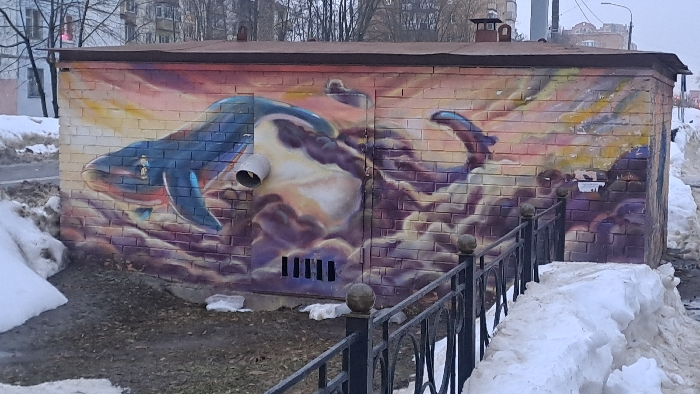 An exterior image taken in Bel'kovo, Russia, displays a single-story brick utility building featuring a prominent mural, observed during what appears to be a post-winter or early spring season. The building has a brown, gently sloped roof and brick walls painted with a vibrant mural depicting a large blue whale amidst swirling purple, yellow, and orange cloud-like forms. A white, cylindrical pipe protrudes from the building's left-center facade, interrupting the mural. Below this, on a central metal access panel, five vertical black vents are present. The ground immediately surrounding the building consists of dark, muddy patches interspersed with irregular accumulations of dirty, melting snow. Large snowdrifts are visible on both the left and right sides of the structure. In the foreground, a section of a black metal fence with decorative spherical finials runs parallel to the building on the right. The background on the left shows bare deciduous trees and a multi-story residential building with a visible red light source. The background on the right includes utility poles, a street light, and additional distant buildings. No human subjects are present in the scene.