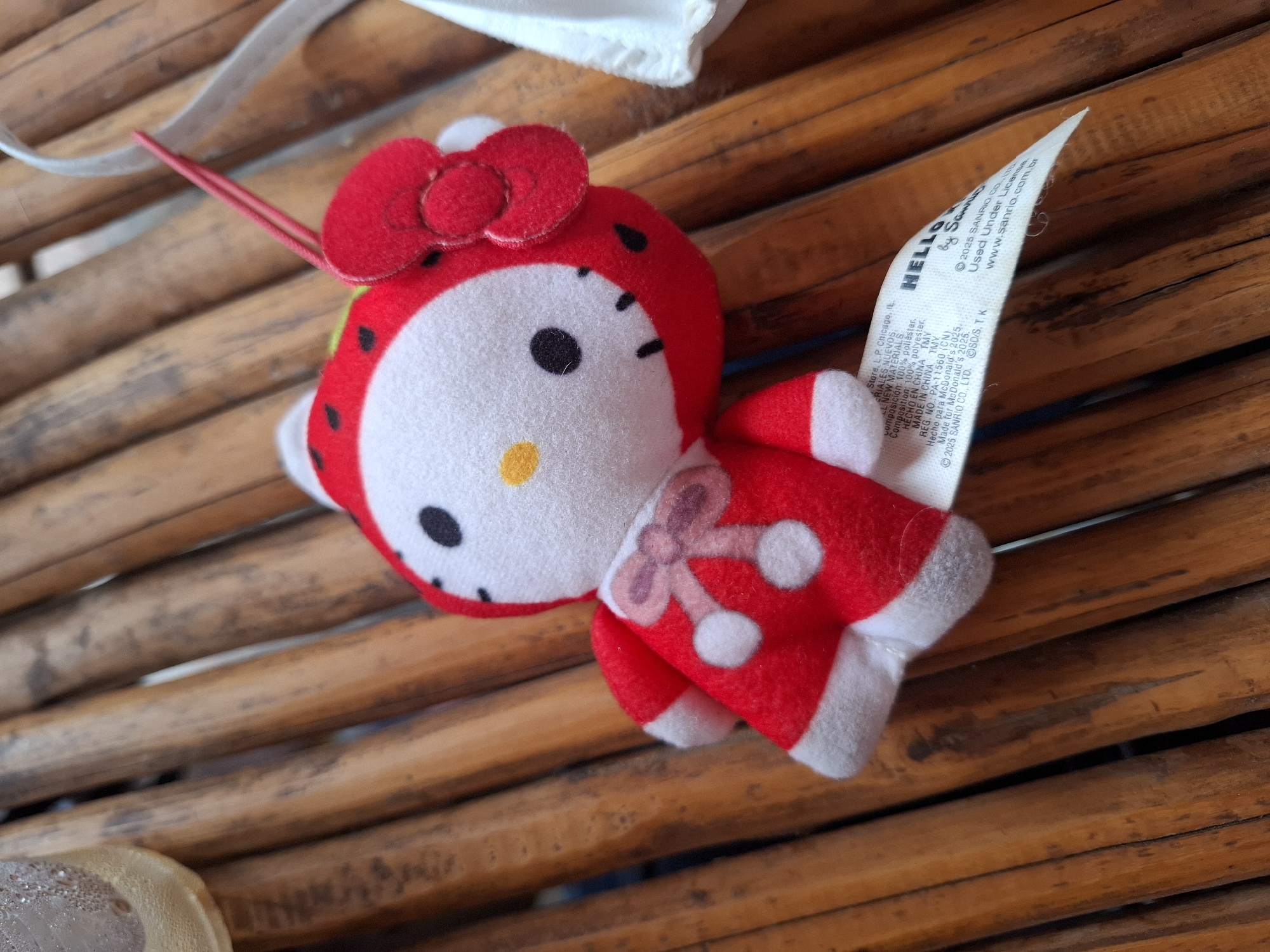 The image depicts a Hello Kitty plush toy lying on a bamboo surface. The toy is red and white, wearing a red dress with white polka dots and a matching red bow. A white tag is attached to the toy, displaying the Sanrio and McDonald's logos. The toy appears to be in a still and static state. The bamboo surface is made up of several parallel wooden slats.
