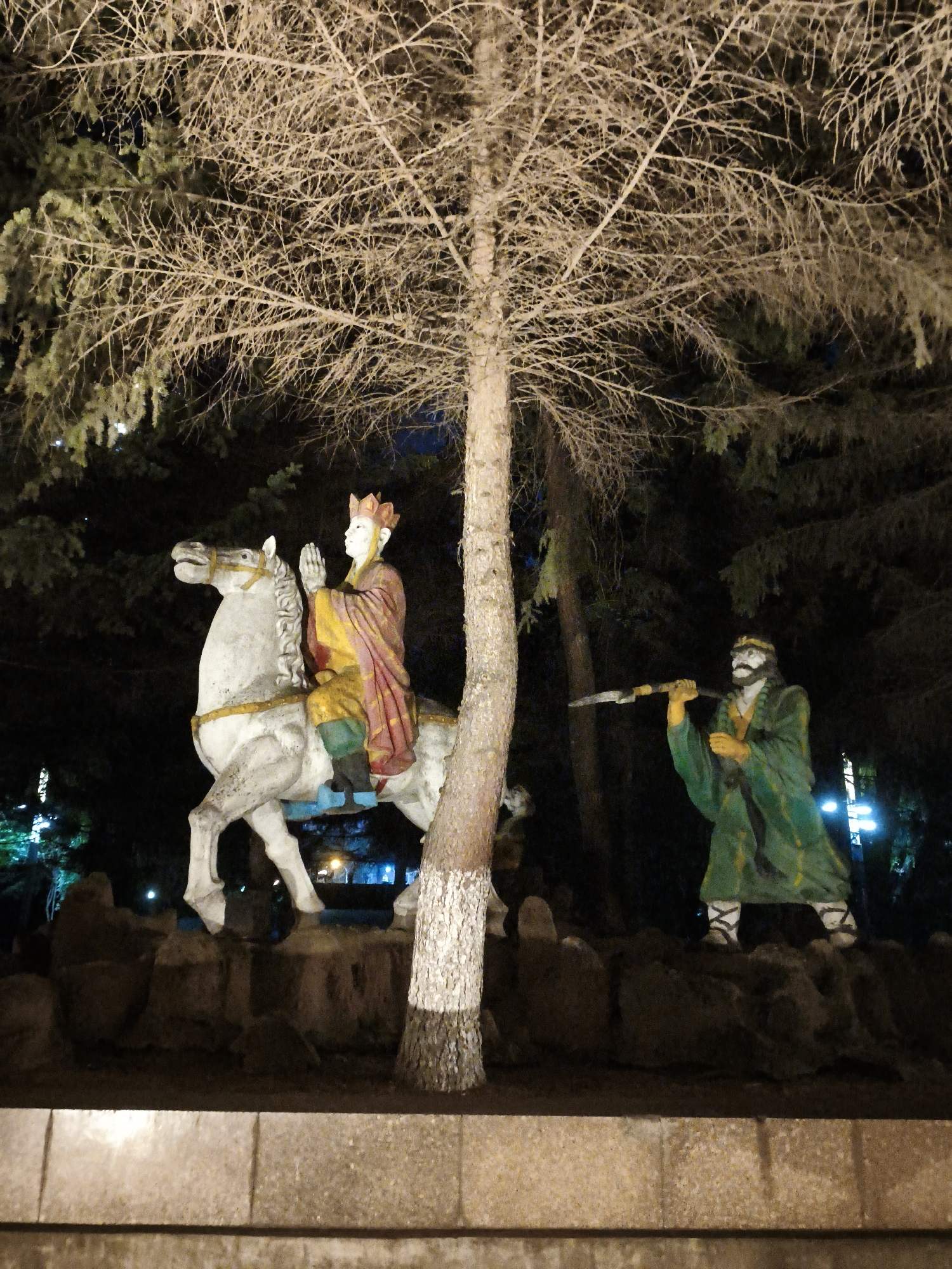 This image captures an outdoor nighttime scene at Dongxing Street, China, featuring two statues and a prominent tree. On the left, a white horse statue is mounted by a figure wearing a red and yellow robe and a crown, with hands clasped in a prayer-like gesture. To the right, another statue depicts a man with a beard, dressed in a green robe, holding a spear-like object. The statues are positioned on a rocky mound, with a large, bare tree standing in the foreground between them, its branches stark against the dark background. The environment is illuminated by artificial light, highlighting the figures and the tree.