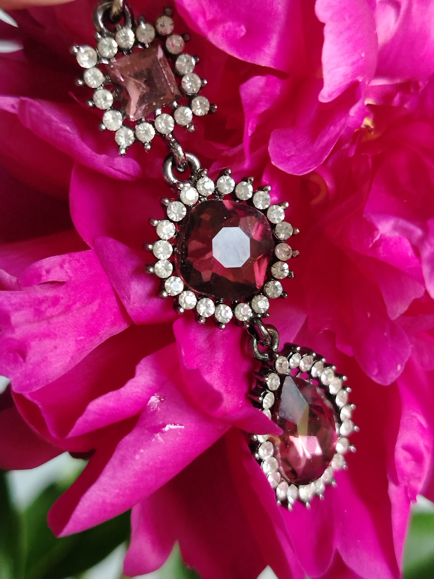 A close-up, high-angle shot captures a piece of ornate jewelry, likely an earring or pendant, resting on the vibrant pink petals of a flower. The jewelry features several faceted gemstones, predominantly a deep pink or ruby red color, surrounded by smaller, clear, diamond-like stones. The metal setting appears to be dark or antiqued silver. The flower's petals are soft and have a velvety texture, with delicate folds and curves. The lighting highlights the sparkle of the gemstones and the lushness of the petals. The background is softly blurred, emphasizing the central subject. There are no people or specific city locations visible in this image.