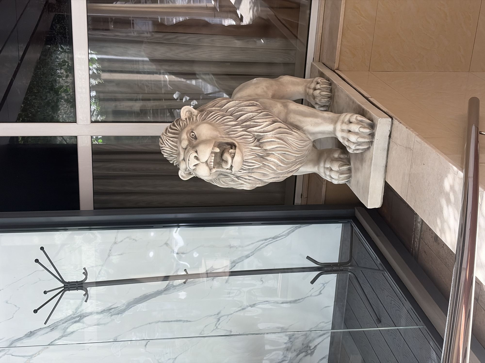 A statue of a lion stands on a stone ledge in front of a building with large windows. The lion statue is light-colored and has an open mouth. Behind the lion, sheer curtains hang in front of the windows. To the left of the lion statue, a coat rack with several hooks is visible through a glass partition. The floor inside the partition appears to be dark wood. The building exterior is tiled in a light tan color. The scene appears to be outdoors, with sunlight casting shadows. This image was taken in the city of Sochi, Russia.