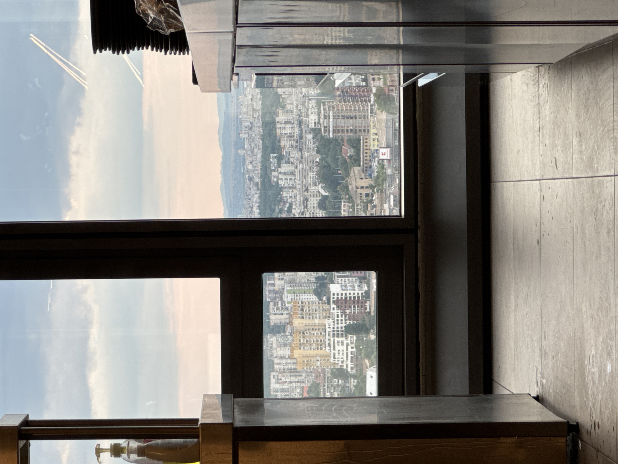 The image shows a view of Sofia, Bulgaria, from a high-rise building. In the foreground, to the left, is a wooden cabinet with a dispenser of liquid soap on top. To the right, there is a metallic cabinet with stacked plates on top. The scene is framed by large windows offering a panoramic view of the city. The windows are divided into sections by dark frames.
The city of Sofia is visible through the windows, with numerous buildings of varying heights and architectural styles. In the middle ground, a dense urban landscape with residential buildings, some of which are yellow and white, is depicted. A red and white "Kaufland" sign is visible on one of the buildings. In the background, rolling hills or mountains can be seen under a cloudy sky. The sky is a pale blue with some white clouds. There are two white trails, possibly from airplanes, visible in the upper right quadrant of the sky. The floor in the foreground is tiled and appears to be slightly wet in places.