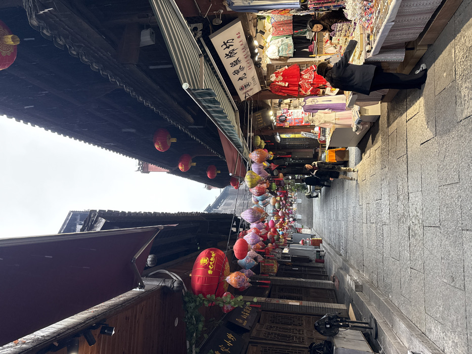 The image displays a narrow, stone-paved commercial street in Pengshui Miao and Tujia Autonomous County, China, under an overcast sky with visible light rain. The street gently inclines uphill, flanked by traditional multi-story buildings characterized by dark wooden facades and tiled roofs. A dense canopy of colorful lanterns, including round red designs and ornate pleated variations, is strung across the street overhead.
On the right side, an open-front stall, identified by a white sign bearing Chinese characters including "九娘服饰坊" (Jiuniang Clothing Workshop), showcases garments such as red traditional dresses and various patterned apparel. Two women are present at this stall: one, wearing a black jacket and pants, examines items on a display table, while another woman, likely a vendor, stands behind the table. Additional commercial stalls are visible further along the street on the right.
The left side of the street is lined with buildings featuring dark wooden exteriors and closed doors. A black floor fan is positioned on the sidewalk in the foreground. In the mid-ground, a man dressed in a dark jacket walks uphill on the stone pavement. Another individual is seated on a structure further along the left side of the street. The stone pavement appears damp.
