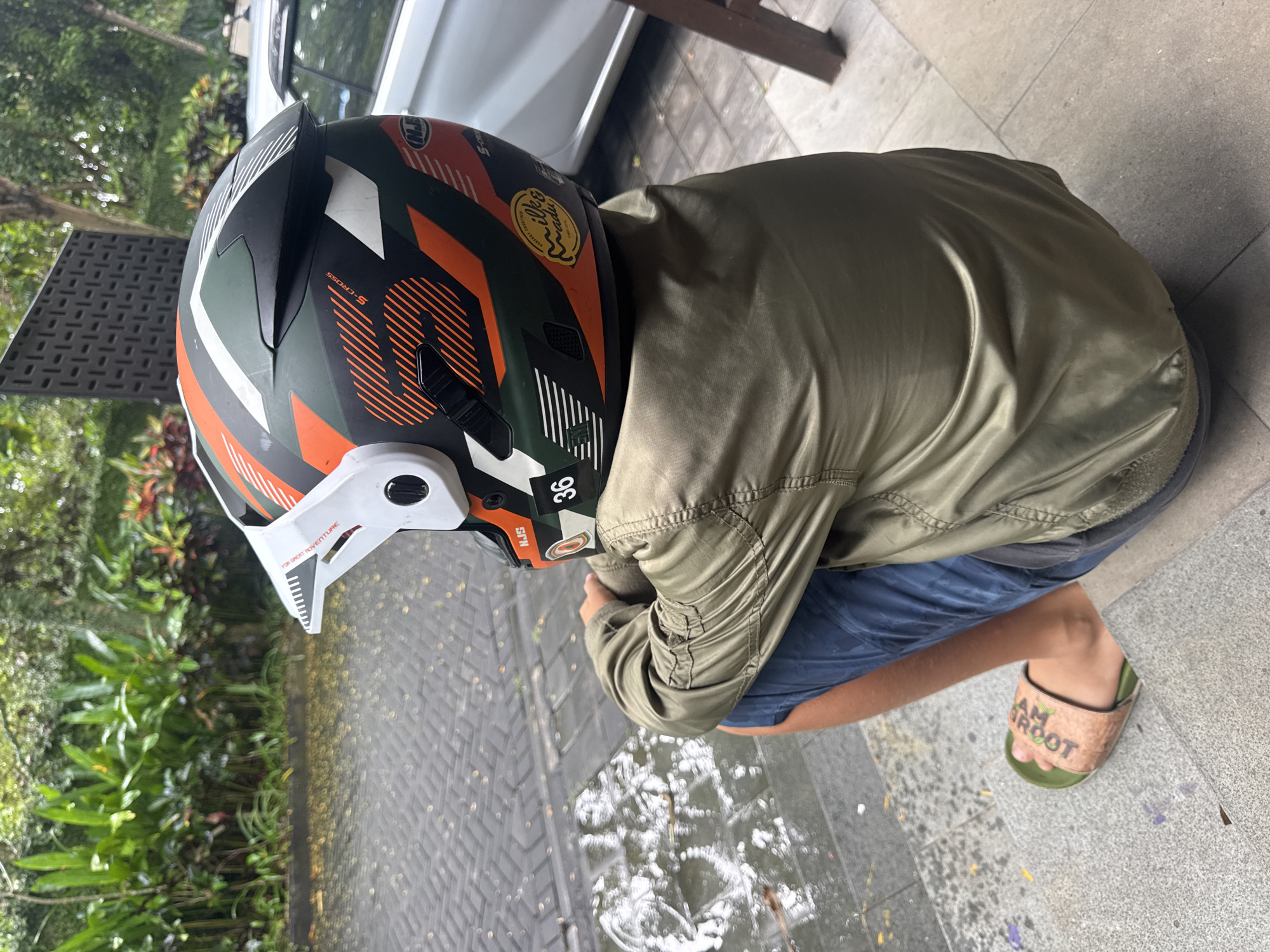 A person wearing a multi-colored helmet, an olive green jacket, blue shorts, and "I am Groot" sandals is crouching on a gray tile surface. The helmet has orange, white, and black markings, with a white visor. The surrounding environment includes greenery, a gray brick-paved area with water puddles, and a section of a white car. The location is Kuta Selatan, Indonesia.