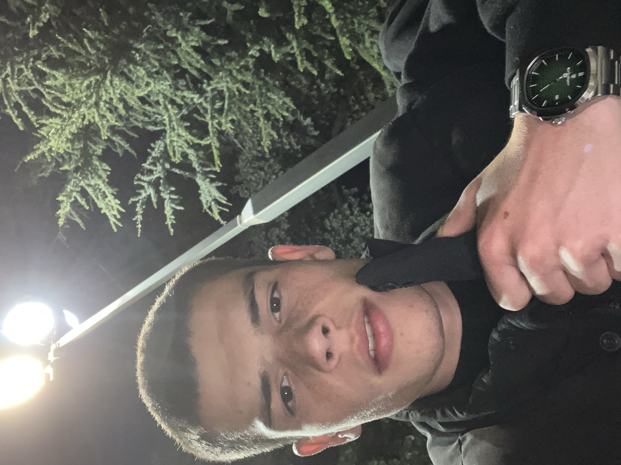 A dimly lit outdoor scene shows a person from a low-angle, close-up perspective. The individual has short, dark hair, light skin, and is wearing dark clothing. A wristwatch is visible on their left wrist. A light source illuminates the person from the bottom-left corner. In the background, there is a light pole and a tree with dense foliage. Specific location information for Karavelovo, Bulgaria, is not discernible from the image.