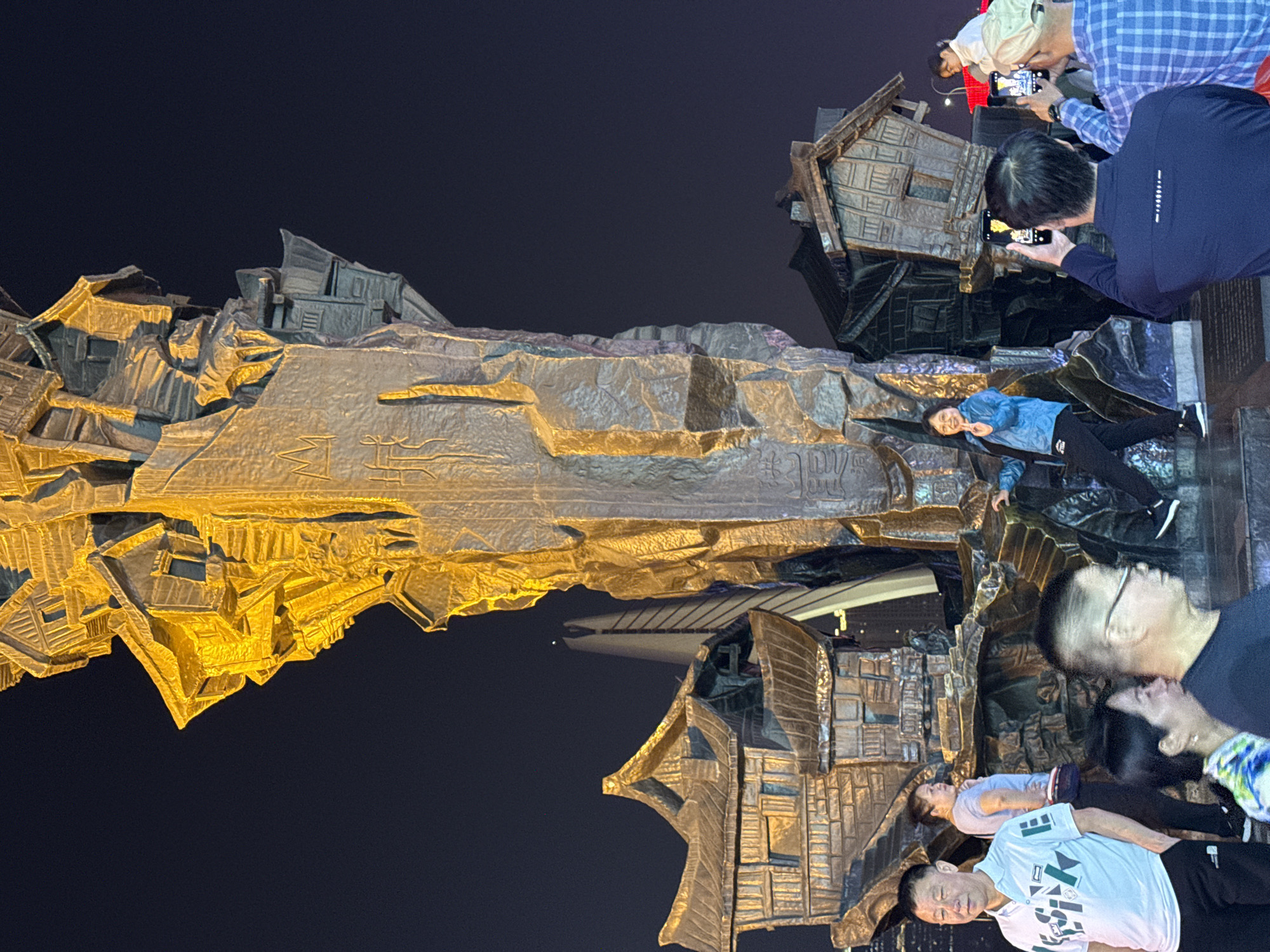A nighttime outdoor scene shows a large, abstract metallic sculpture in Yuzhong District, China, illuminated from below. The sculpture appears to depict a landscape with stylized buildings or houses integrated into the terrain. Several people are interacting with the sculpture. Two people are taking pictures of the sculpture. A woman in a blue jacket and black pants is perched atop a section of the sculpture. Two other people are posing at the base of the sculpture. The backdrop is a dark night sky.