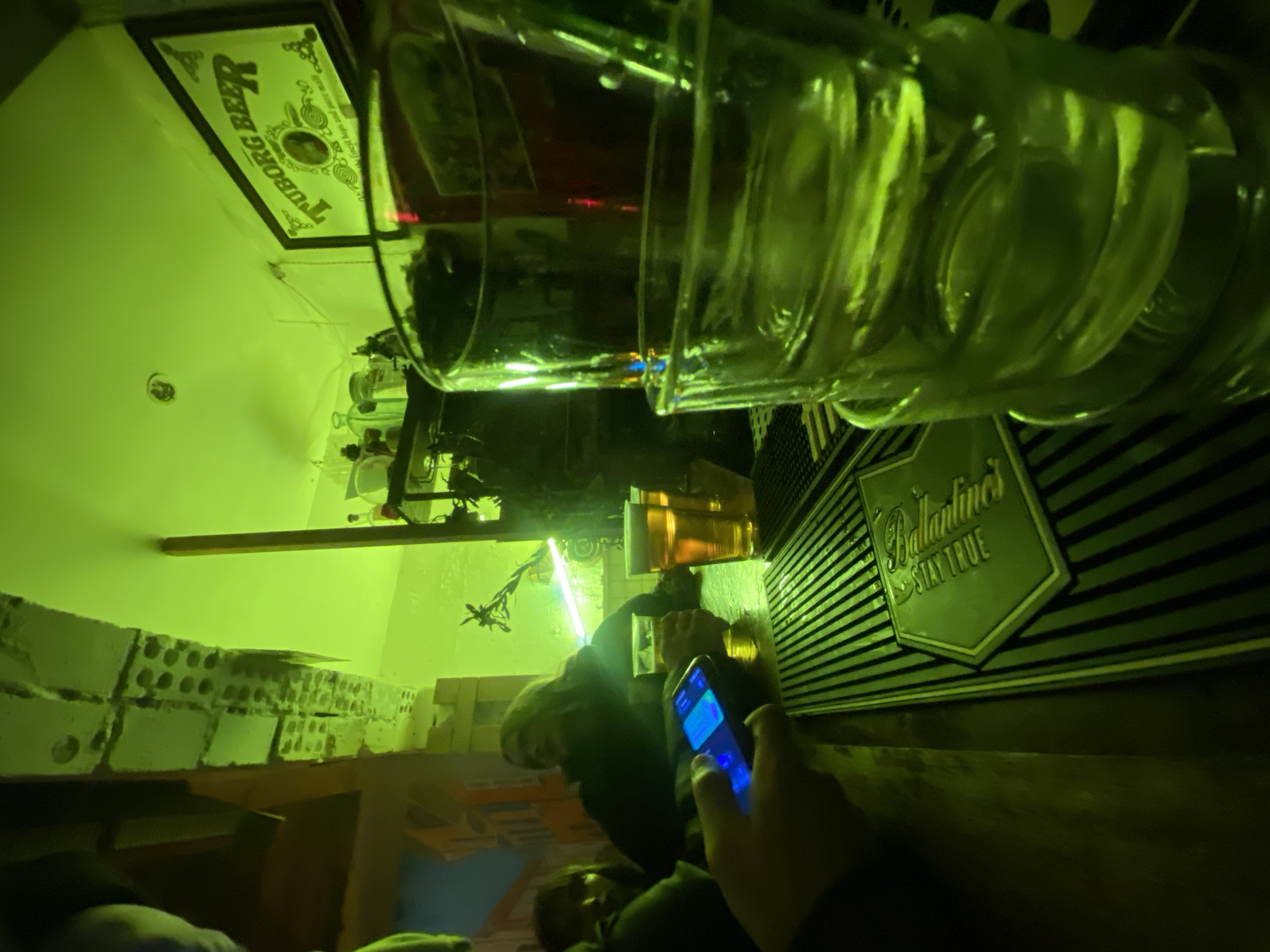 This image captures an indoor scene, likely a bar or pub in Moscow, Russia, characterized by a strong green ambient light. The perspective is from a patron seated at the bar, looking across the counter.
In the immediate foreground, a large, clear glass, possibly a beer mug, lies horizontally on the bar counter. Its surface reflects the green light, creating highlights. To the right, a hand is visible, holding a smartphone with a brightly lit blue screen, resting on the bar.
The bar counter features a dark, ribbed or textured surface. On it, a metallic-looking sign with the brand "Ballantine's STAY TRUE" is prominently displayed. Further along the counter, another glass containing a golden liquid can be seen, partially illuminated. Behind the bar, several bottles are visible, some reflecting light.
In the mid-ground, several individuals are present, mostly obscured or seen from behind. One person with dark hair is seated or standing near the bar, facing away from the camera. The dark setting and artificial lighting suggest the image was taken during the evening or night.
The background elements include a wall on the left constructed from light-colored bricks or cinder blocks. On the upper left wall, a framed sign for "TUBORG BEER" is mounted. A light fixture is visible on the ceiling or upper wall, contributing to the green glow. The overall atmosphere is dim and intimate, typical of a late-night social setting. The scene depicts people enjoying drinks and socializing in a relaxed environment.