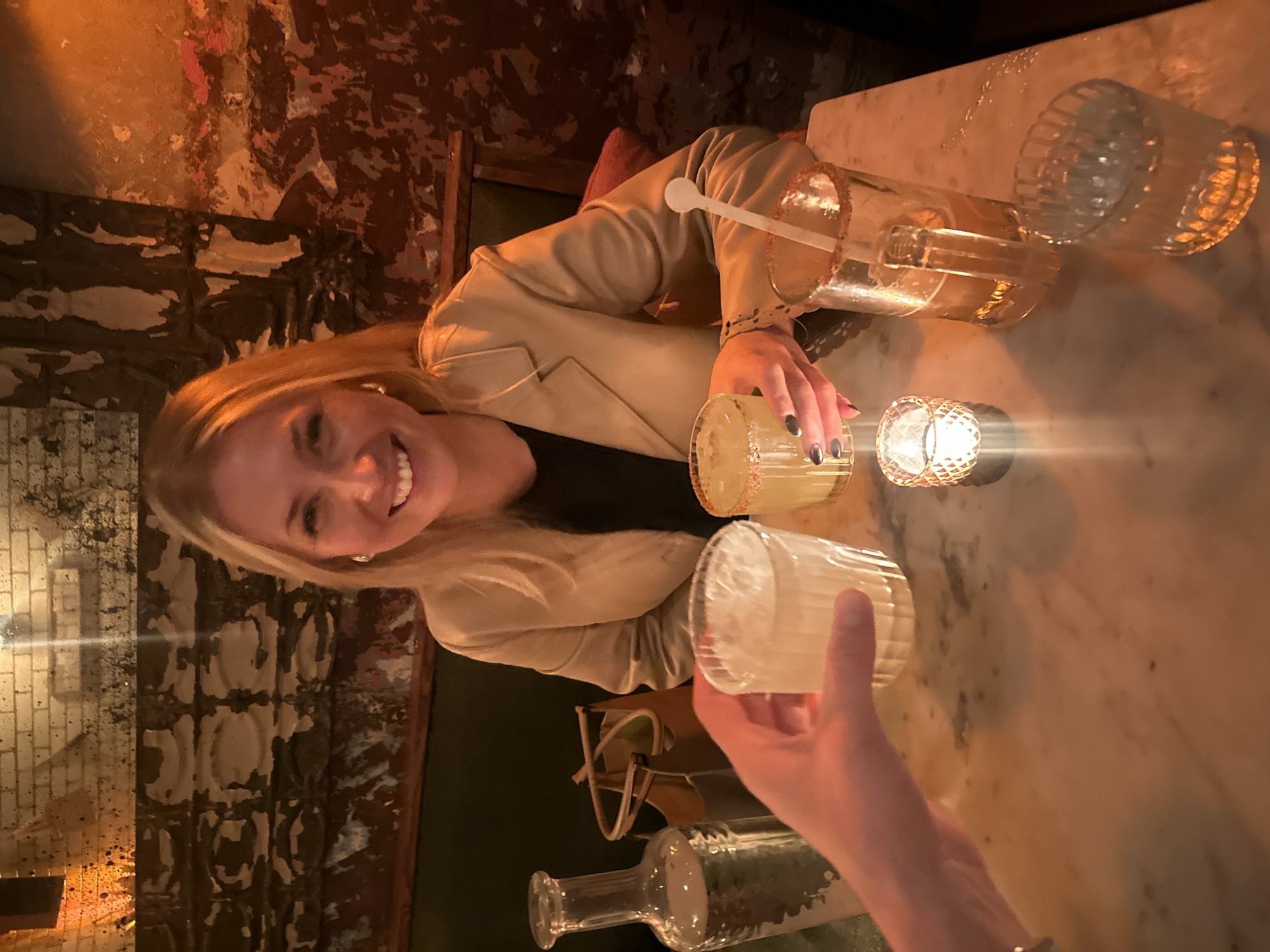 Here is a detailed description of the image:
The image shows a woman with blonde hair smiling at the camera while sitting at a marble-topped table. She is wearing a light tan blazer over a dark top. On the table, there are several glasses, including a large pitcher filled with a light-colored liquid and a spoon inside, as well as other glasses containing liquids. There is also a decorative candle holder with a lit candle. A hand reaches into the frame, holding a glass. The background features a wall with textured, distressed paintwork in shades of brown, red, and beige. The lighting appears to be dim, creating a warm and intimate atmosphere.
Based on the style and the general ambiance, the image could have been taken in London, United Kingdom, known for its upscale bars and restaurants. However, without specific visual clues, it's impossible to pinpoint the exact location within the city.