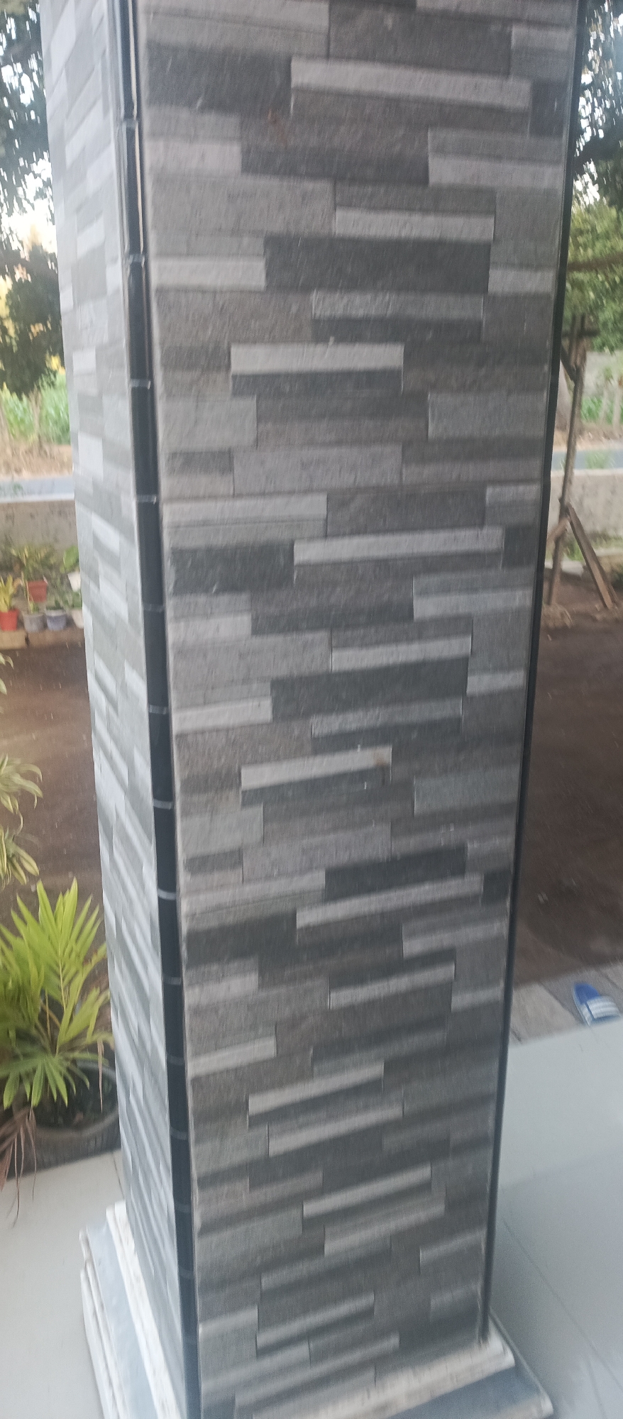 The image shows a close-up of a pillar covered with rectangular stone tiles in various shades of gray. The tiles are laid in a staggered pattern, creating a textured surface. The pillar is vertical, with a black vertical trim running along its left edge. In the background, a lush green plant in a dark pot is visible to the left, and out-of-focus foliage and trees suggest an outdoor environment. A hint of a white structure and a light blue object, possibly a slipper, are present on the right side. The overall scene is an outdoor setting, likely a residential or public area in Jatisari, Indonesia.