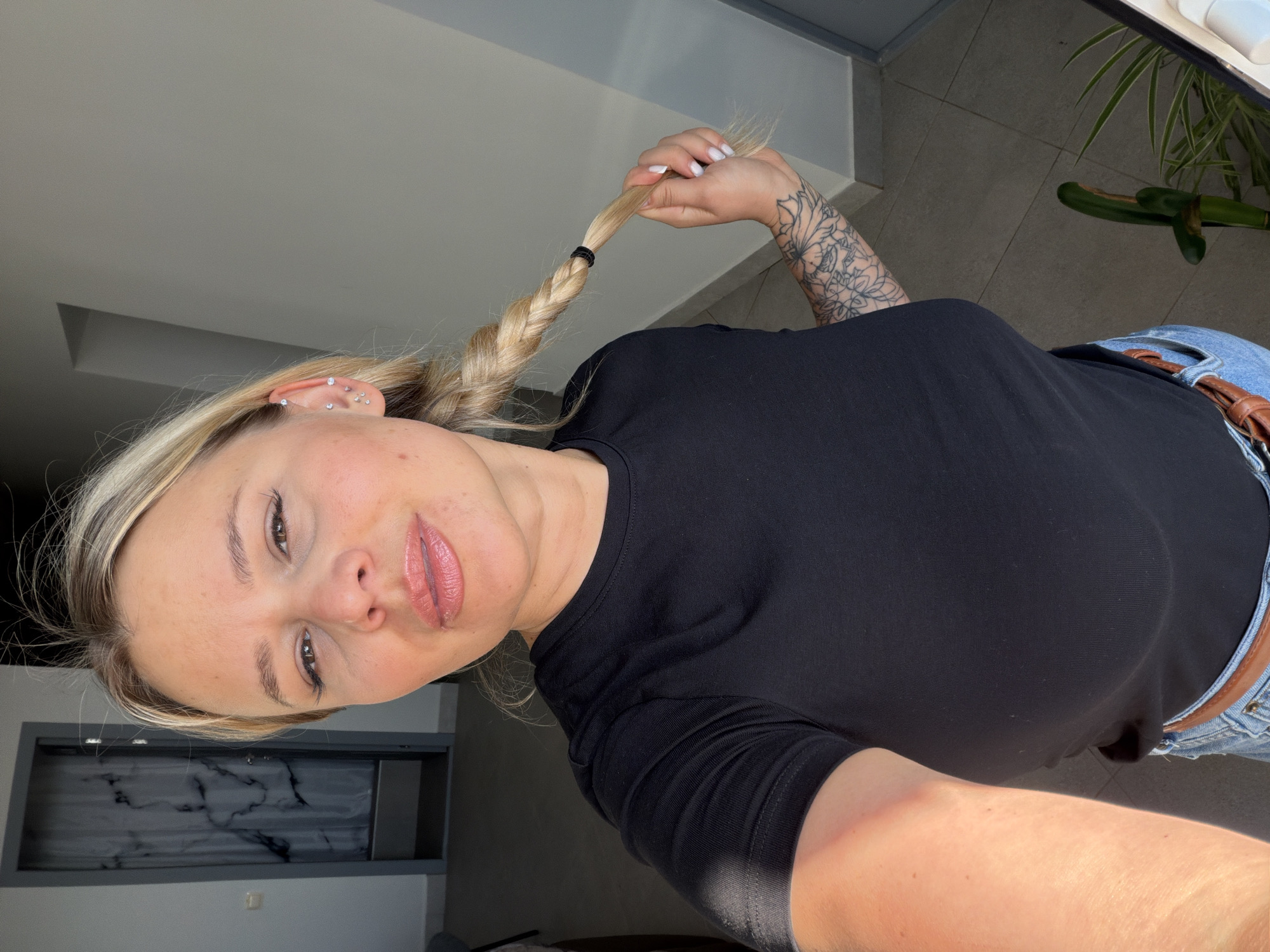 A blonde woman with fair skin and light brown eyes is holding the end of her blonde braid in her right hand. She is wearing a black t-shirt, blue jeans with a brown belt, and has a tattoo on her right forearm. She has multiple piercings in her left ear. The background shows a room with a gray tiled floor, a white wall with a door, a plant, and a gray cabinet. The photo was likely taken indoors. Sarapul, Russia is not evident in this image.