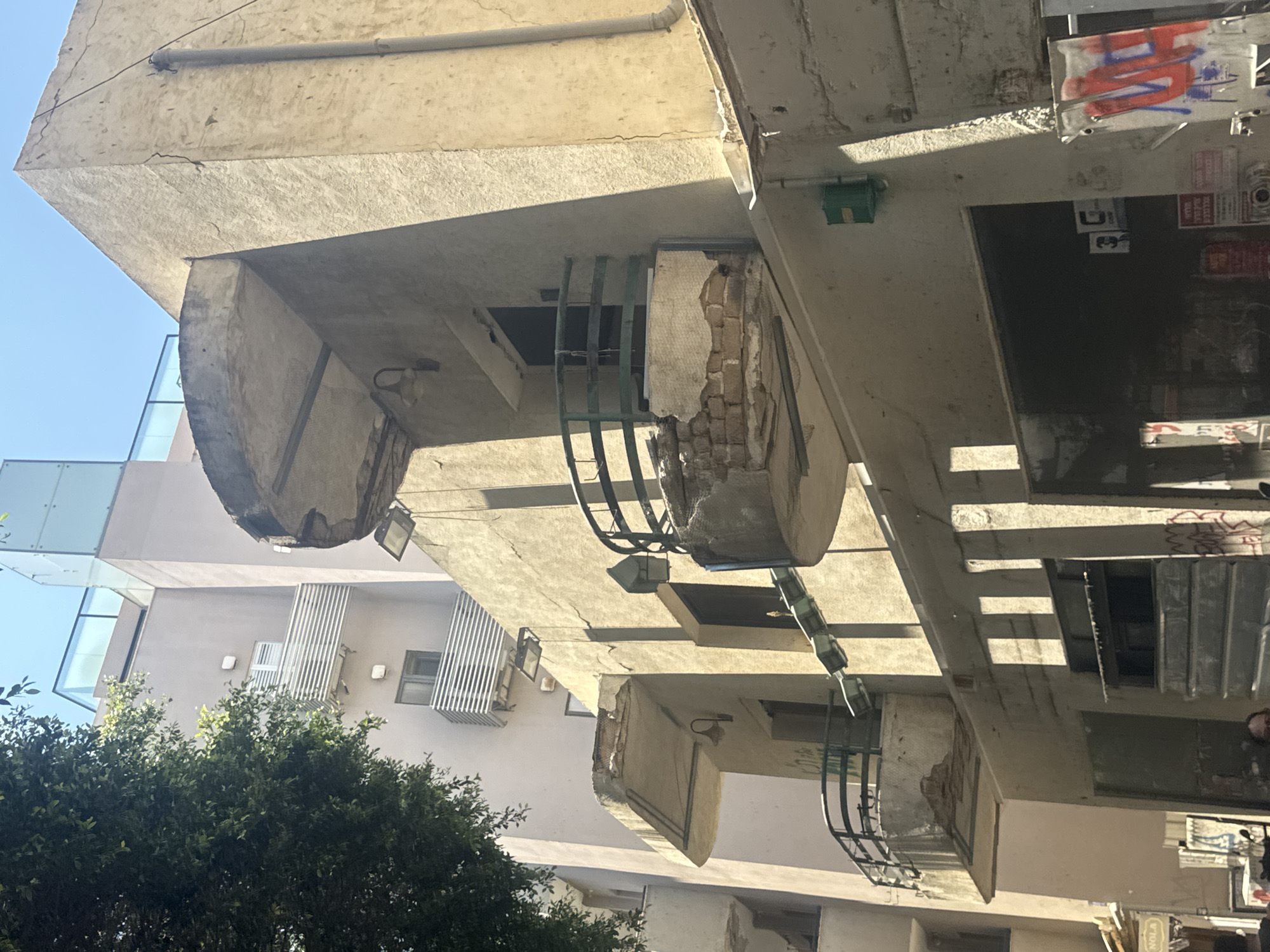 This is a low-angle, daytime shot of the exterior of a building in Tel-Aviv, Israel. The building has overhanging concrete balconies with metal railings. The balconies show signs of age and disrepair with exposed brick and crumbling concrete. Sunlight casts shadows across the building. In the lower left, there is a green tree. In the upper left, a building with large glass windows is visible. Graffiti is visible on the building.