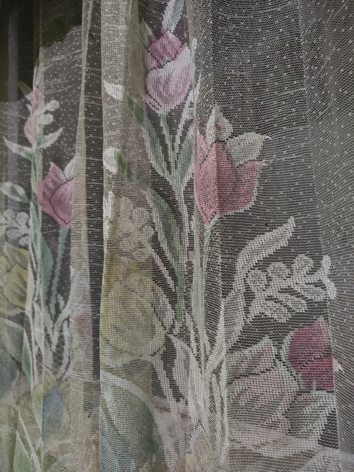 A close-up forensic examination of a sheer, light-colored textile curtain, located within a structure in Astanajapura, Indonesia. The curtain features a repeating woven or embroidered floral pattern, primarily depicting stylized pink/mauve and green flowers, leaves, and stems against a darker, indeterminate background visible through the translucent material. The fabric exhibits a fine mesh or lace-like weave with a subtle dotted texture. The curtain appears slightly draped or folded, creating vertical lines within the composition. No individuals, actions, or additional objects are visible within the frame.