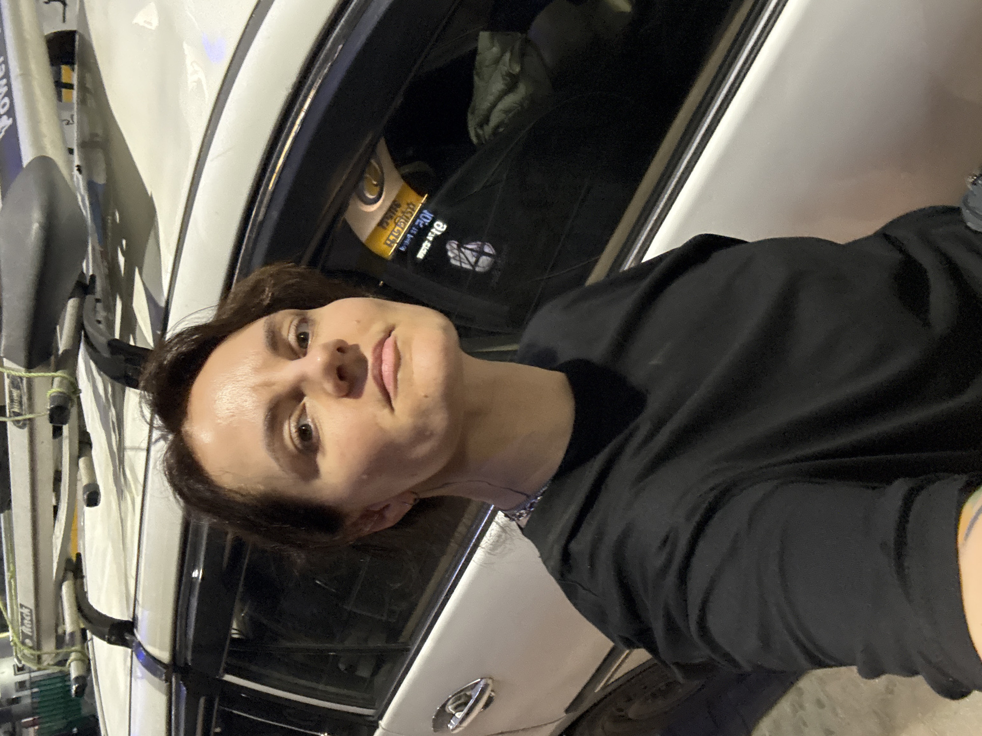 This media file captures a selfie of a person with short, dark hair and a neutral expression, wearing a black long-sleeved shirt. The lighting indicates it is nighttime or late evening, with artificial light illuminating the subject and a white car in the background.
The person is standing next to the white car, which is equipped with a roof rack. On the side of the roof rack, the word "Power" is visible in blue text. Through the car's window, parts of the dark interior and a grey item on the back seat are discernible. A yellow sticker is affixed to the inside of the car window. This sticker displays text in an Indian script, including the numbers "०७५१०" (07510), and below it, the text "B Bicycle" alongside a white symbolic logo that resembles a fist. A chrome door handle is also visible on the car.
The scene suggests a stop or a moment during travel, possibly in Bagdogra, India, given the location context provided and the Indian script on the car sticker. The presence of the roof rack implies the car is used for transporting equipment, potentially for outdoor activities like cycling.