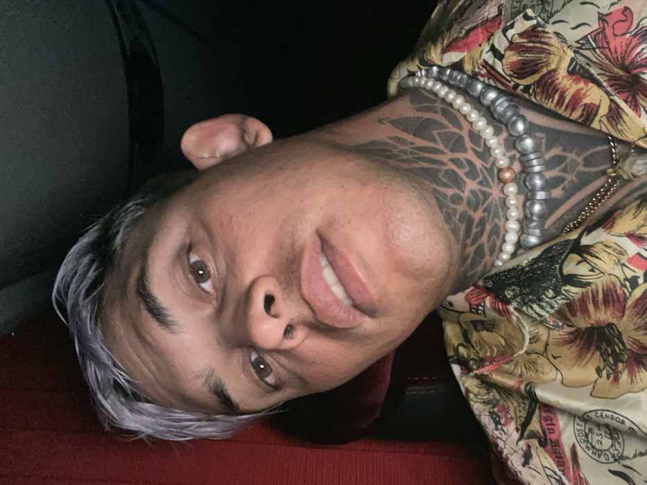 A person with short, gray-tipped hair lies at an angle, smiling slightly. They have brown eyes and a noticeable geometric tattoo covering much of their neck. Several strands of pearls and silver beads adorn their neck. They are wearing a patterned shirt with floral and text-based designs. A dark surface and a red textured material are visible in the background.