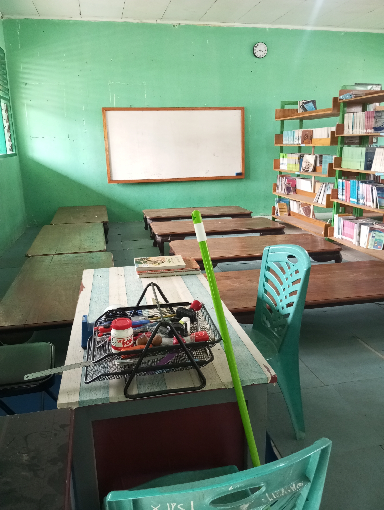The image depicts a classroom or library setting, likely at a school in Pematang Reba, Indonesia, given the context of the provided files. The room is painted a light green, with white ceiling tiles. A large, empty whiteboard with a wooden frame hangs on the wall to the left. In the center of the room are several low, wooden tables, some arranged in rows. To the right, a tall, green metal bookshelf is filled with books of various sizes and colors. A blue plastic chair is positioned near the bookshelf. In the foreground, a smaller table has a metal organizer holding various school supplies, including a red jar labeled "lem FOX." A long, bright green cleaning mop leans against the table. The lighting suggests it is daytime, with sunlight casting shadows through a window on the left. The floor appears to be tiled or linoleum. The overall impression is a functional, perhaps slightly aged, educational space.