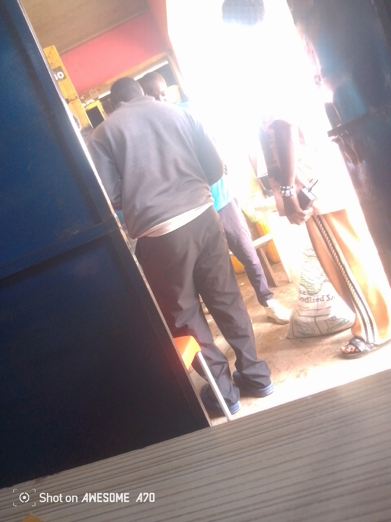 The media file shows a scene with several people in what appears to be an indoor setting, possibly a market or shop. The lighting is very bright, suggesting it could be daytime or that artificial lights are in use.
In the foreground, a person is seen from behind, wearing a grey sweatshirt and dark pants. This individual is standing next to a blue structure. To the right of this person, another individual is visible, also from behind, wearing light-colored pants and a bright white shirt, with their arm adorned with bracelets. This person is holding a dark object. Further to the right, a person is wearing light-colored pants and sandals. A large bag, possibly containing a powdery substance like salt, is visible on the ground in front of this person, with text that partially reads "ODIZED Sa". Other people are partially visible in the background, one wearing a light blue shirt and dark pants, and another figure further back. The overall scene seems to depict a moment of everyday activity, perhaps shopping or waiting. The bright lighting and the casual attire of the individuals suggest a warm environment. There is also text visible at the bottom of the image that reads "Shot on AWESOME A70".