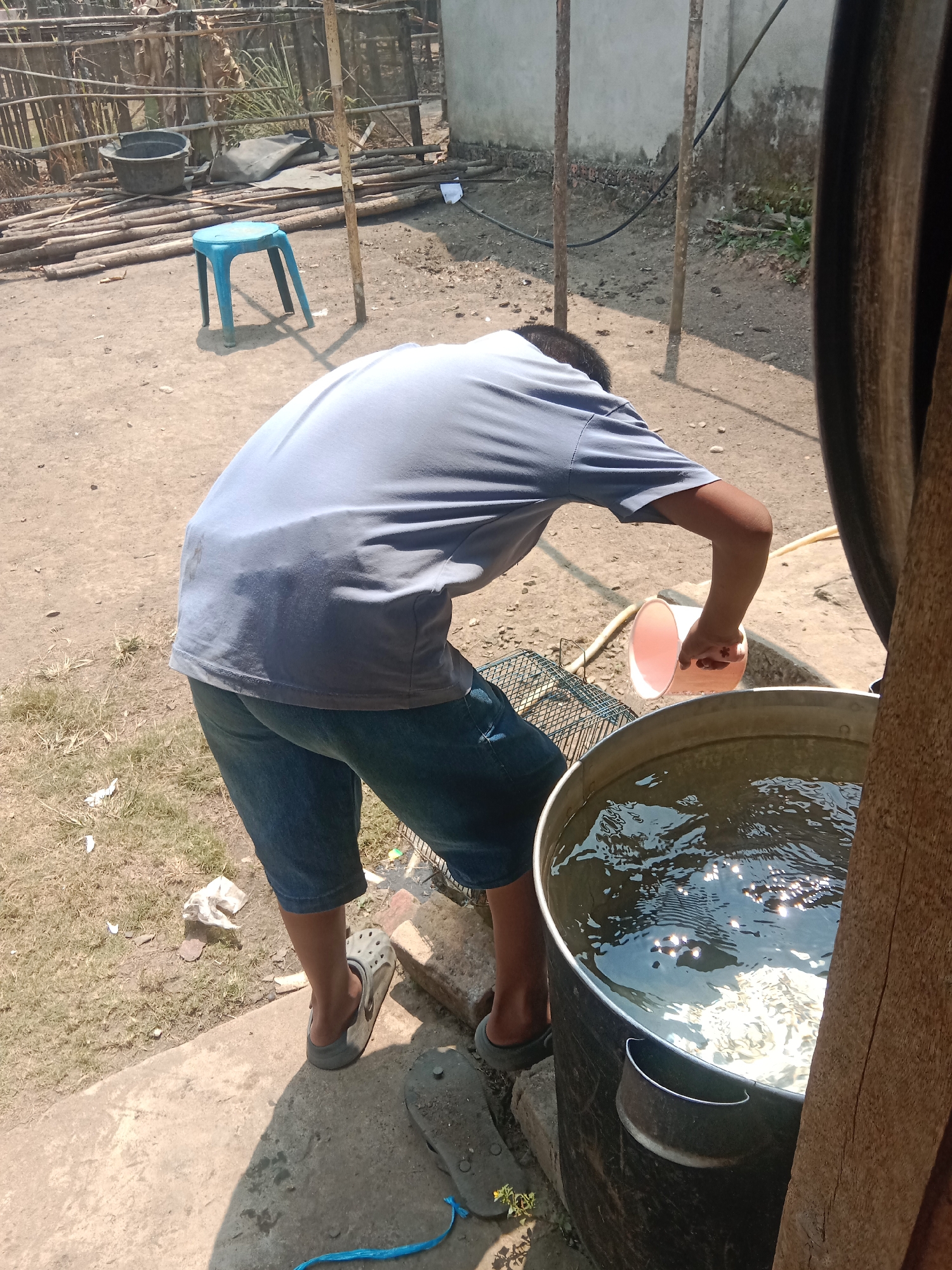 In Paya Besar, Indonesia, a person is depicted pouring water from a pink plastic cup into a large metal container filled with water. The individual is wearing a grey t-shirt and blue denim shorts, along with grey Croc-style sandals. The scene takes place outdoors on a dirt and concrete ground, with a backdrop of wooden structures and fencing. A small blue stool is visible in the background, as are various logs and a large black tub. The sun is shining, casting shadows on the ground.