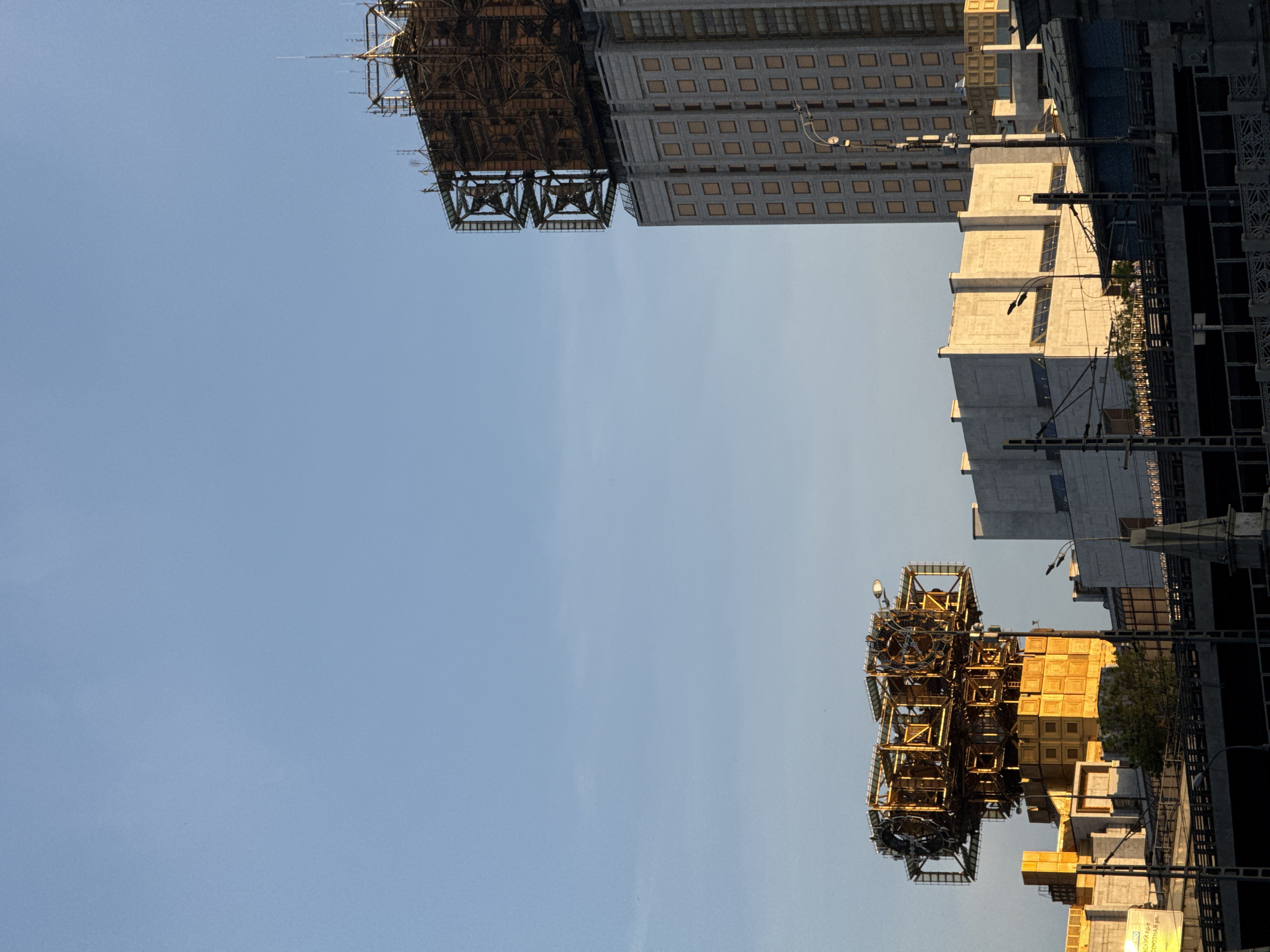 The image shows a view of buildings and structures in Moscow, Russia, from a low angle. The lower portion of the image includes complex, cage-like metal structures, possibly scaffolding or construction elements, with visible light fixtures. One of the buildings has a grid pattern of recessed windows. In the upper portion of the image, there is an additional construction crane and a completed building. The sky is clear and pale blue. The buildings appear to be modern, possibly in a commercial or residential area undergoing development.