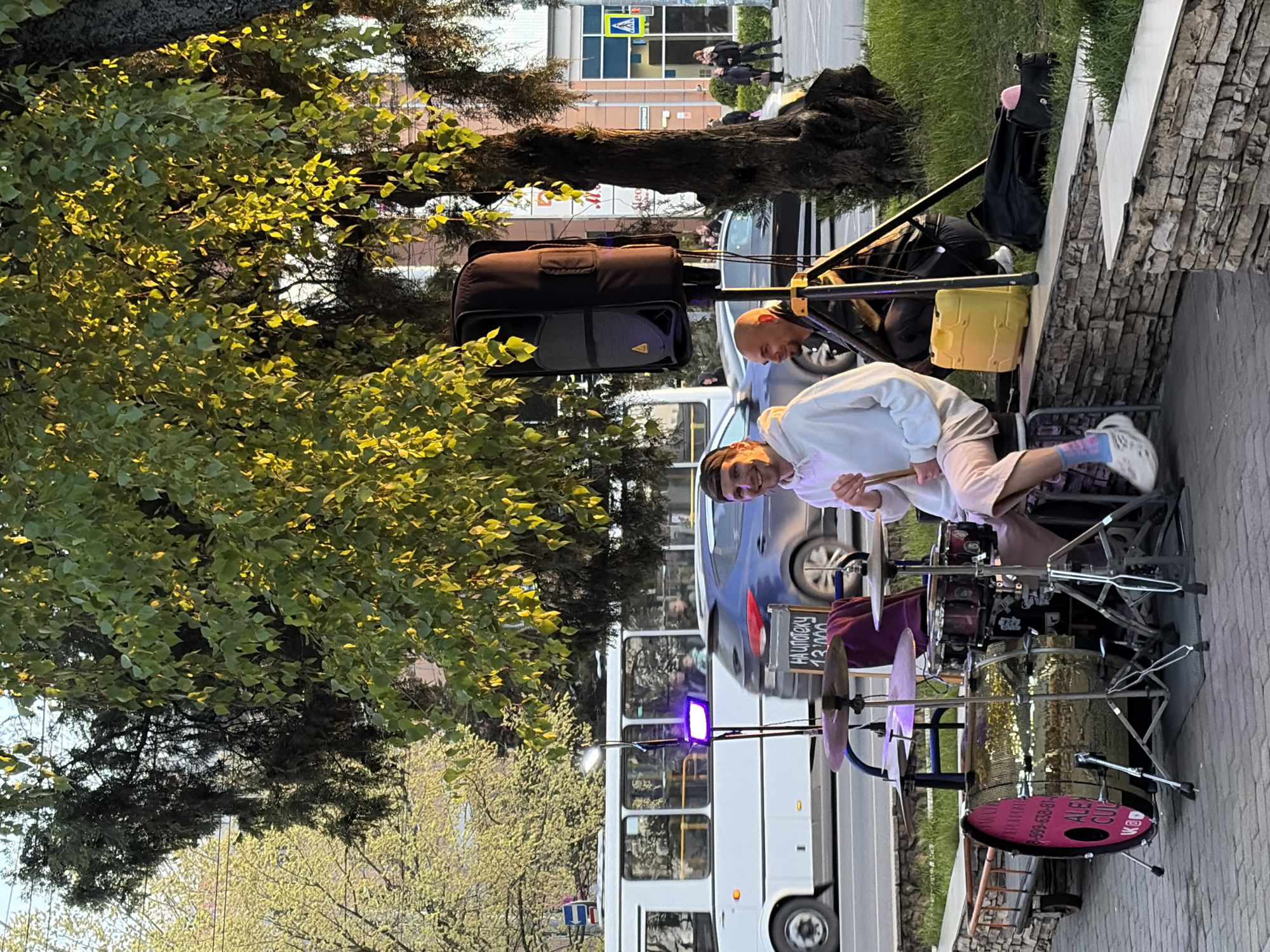 The image captures an outdoor street performance by a drummer in Krasnodar, Russia, during what appears to be late afternoon or early evening, indicated by the golden light illuminating the foliage. The sky is clear, suggesting fair weather.
In the foreground, a young man, smiling and wearing a white hoodie and light-colored shorts, is seated at a drum kit, holding drumsticks as if performing. The drum kit features a striking gold glitter finish on the bass drum. The front head of the bass drum is pink and displays social media logos (YouTube and VK) along with partially visible text, including "ALEX OUI" and a phone number.
To the left of the drummer, a bald man is visible, likely assisting with the performance, possibly as a sound engineer, positioned near a large black speaker on a stand. A yellow bucket, possibly for tips, is placed near the speaker stand. Other equipment includes microphone stands and a purple light illuminating the area around the drums. A small blackboard sign, partly visible near the drum kit, reads "НАЛОЖКУ 13,000".
The scene is set on a paved sidewalk next to a stone wall. In the background, a white public bus and a blue car are moving on a road. Further back, buildings and more pedestrians are visible, including a pedestrian crossing sign. Lush green trees, some with light green spring foliage, frame the scene, their leaves reflecting the golden sunlight. The atmosphere appears lively and engaging, typical of street entertainment.