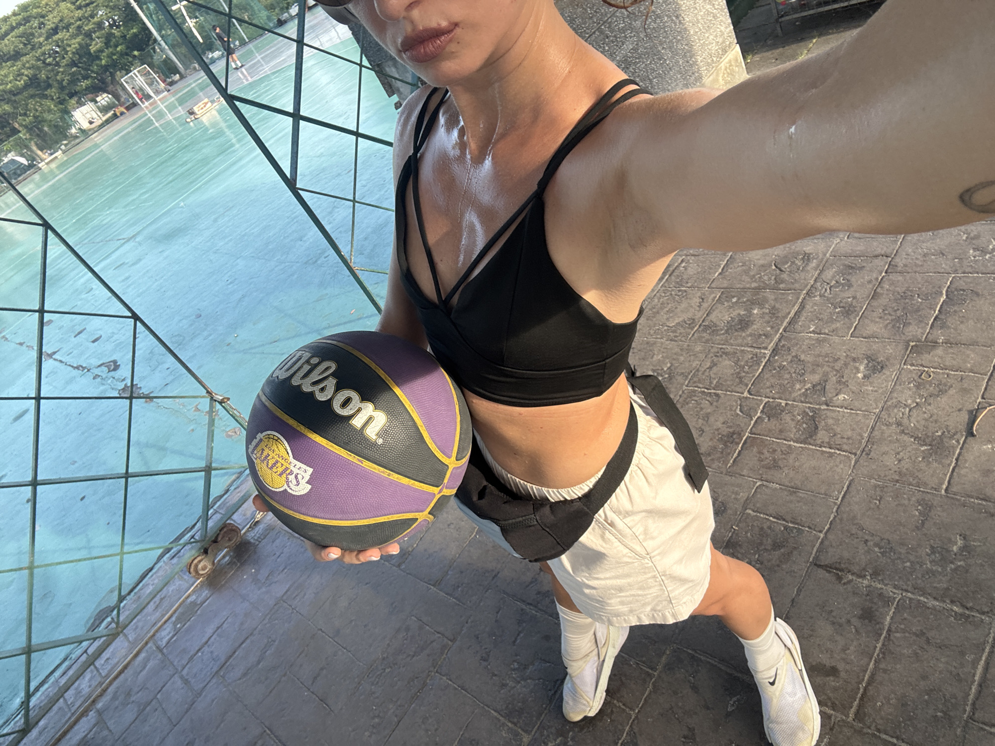In Ko Samui District, Thailand, a person is holding a purple and yellow Los Angeles Lakers basketball on a paved area next to a teal-colored court enclosed by a green net. The person, wearing a black sports bra and white shorts, appears sweaty, suggesting physical activity. The environment is outdoors, with sunlight casting shadows. A portion of a building and trees are visible in the background.