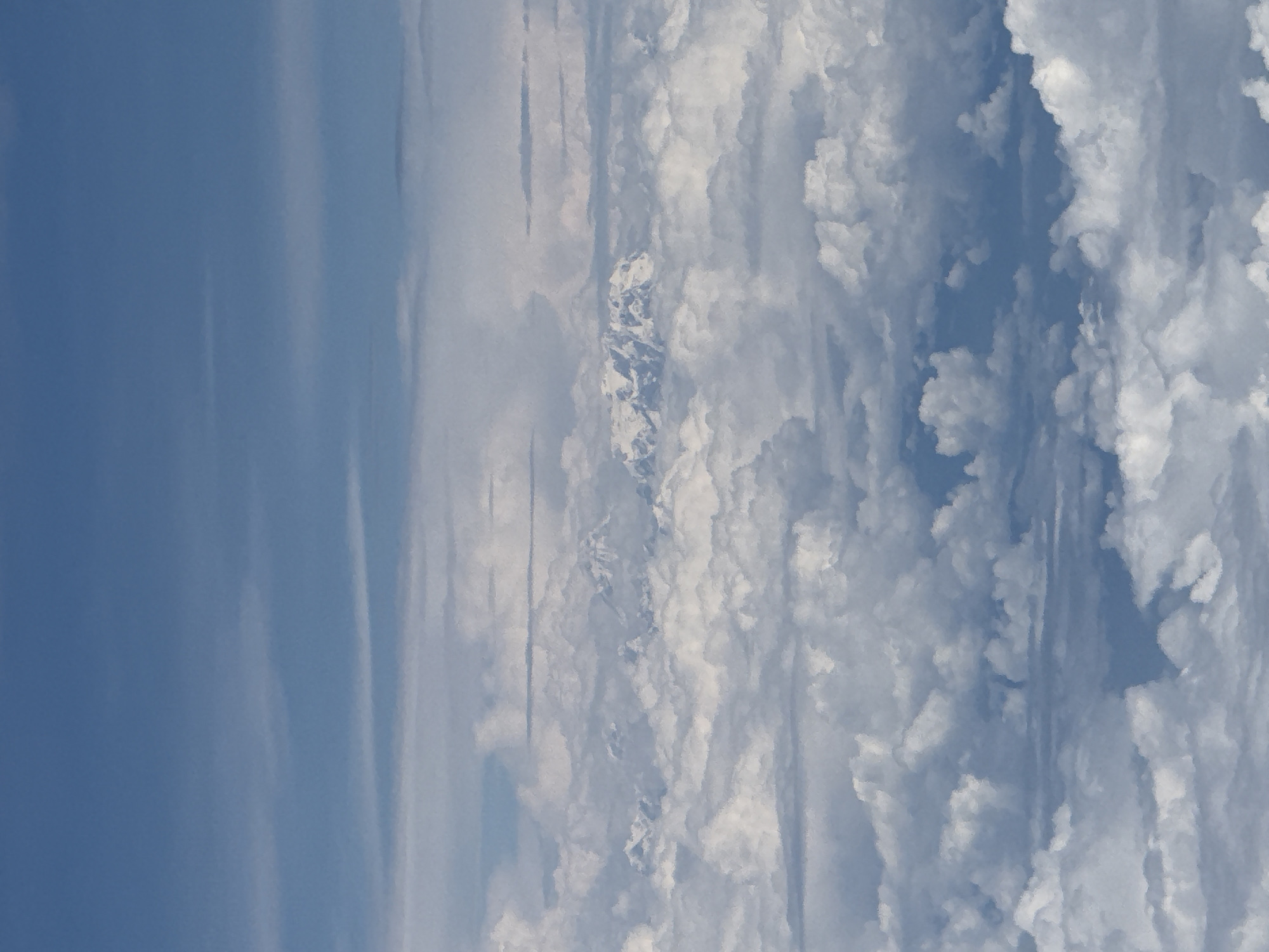 The image displays a view from above the clouds, with the blue sky visible in the upper portion. Several layers of clouds are present, with some appearing more dense and fluffy than others. Peeking through the clouds in the middle distance are snow-capped mountain peaks. There is no discernible indication of the city of Saharanpur, India, nor are there any people, subjects, objects, or actions depicted. The scene is purely atmospheric.