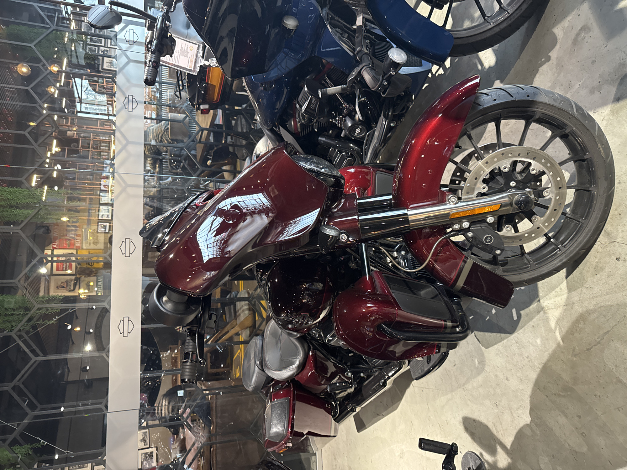 The image shows two Harley Davidson motorcycles indoors. One is dark blue and mostly out of view, while the other is a shiny burgundy color and occupies the main focus. The burgundy motorcycle's front wheel and body are prominently displayed, revealing its brake disc, fender, and tank design featuring a reflective emblem.
Behind the motorcycles is a transparent partition with a honeycomb pattern, showcasing a view of a room with various framed pictures and other decorative elements, suggesting a showroom or dealership environment. Reflections and lighting indicate an interior setting with artificial illumination. The concrete floor is visible in the foreground, and a tool is on the floor next to the burgundy motorcycle. The location is most likely not Saint Petersburg, Russia.