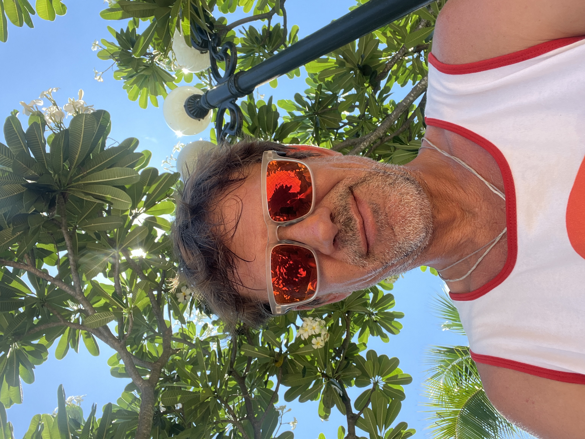 A low-angle, close-up outdoor selfie shows a middle-aged man with short, dark, wavy hair and a graying beard. He is wearing clear-framed sunglasses with bright red lenses that reflect the surrounding environment, a white tank top with red trim, and a thin silver chain. The man is smiling slightly as he looks at the camera. Behind him, lush green foliage and white flowers from a frangipani tree are visible against a bright blue sky. A black lamppost with two illuminated globes stands to the right of the man, partially obscured by the tree. Palm fronds can be seen in the lower left corner of the frame. The lighting suggests it is daytime. The scene appears to be set in a tropical or subtropical outdoor location, possibly a resort or garden area.