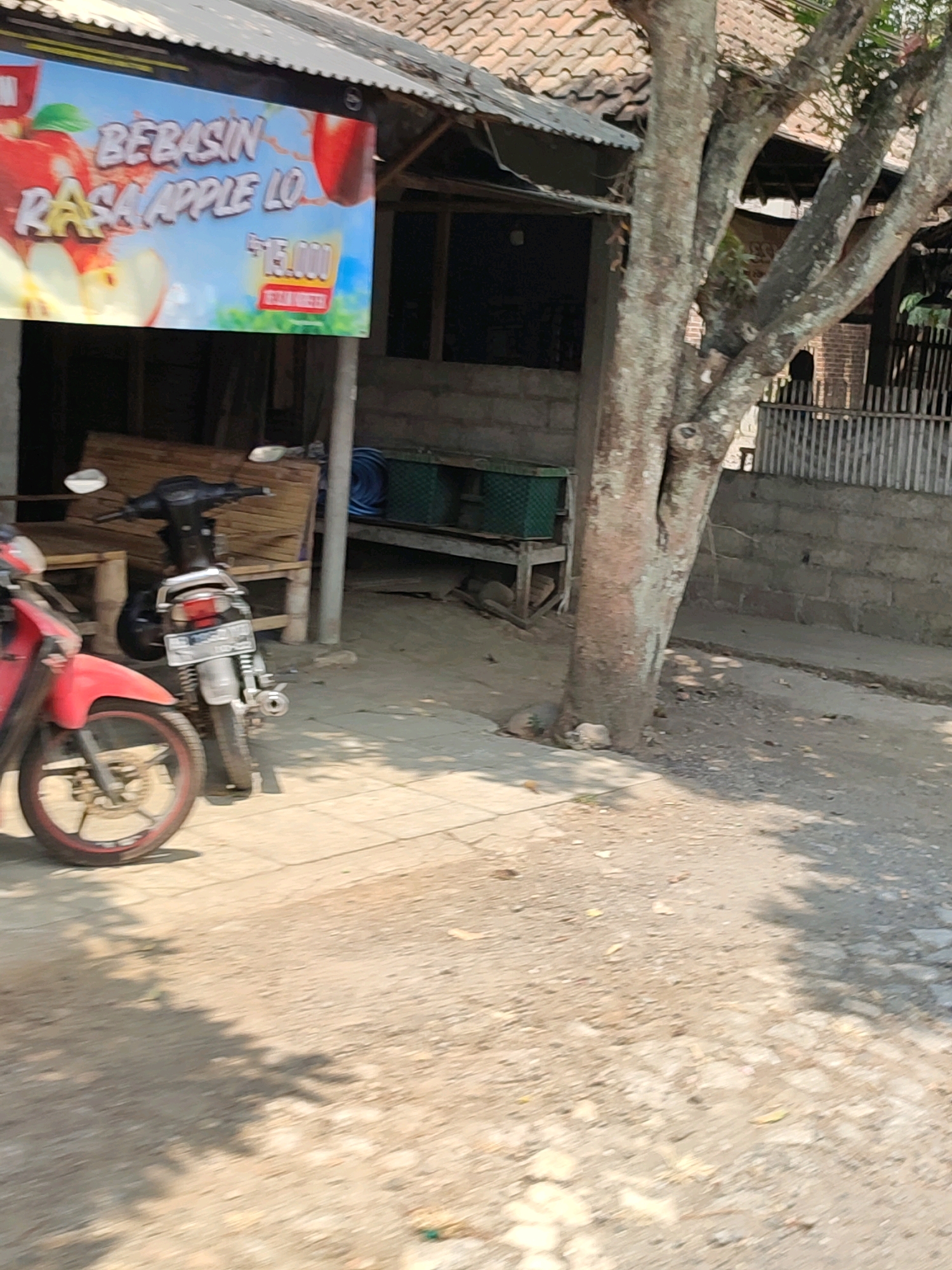 The image depicts a street-side scene in Tanggulrejo, Indonesia, featuring a stall with a banner advertising "BEBASIN RASA APPLE LO" with a price of "Rp. 15.000". Two motorcycles are parked in front of the stall, one red and partially visible, and a silver motorcycle to its right. Behind the motorcycles, there is a wooden structure that appears to be part of the stall or an adjacent building. Further back, a large tree trunk dominates the right side of the frame, with its branches extending upwards. The ground is a mix of dirt and paved surfaces. In the background, a stone wall and a wooden fence are visible. The overall impression is of a local market or food stall setting.