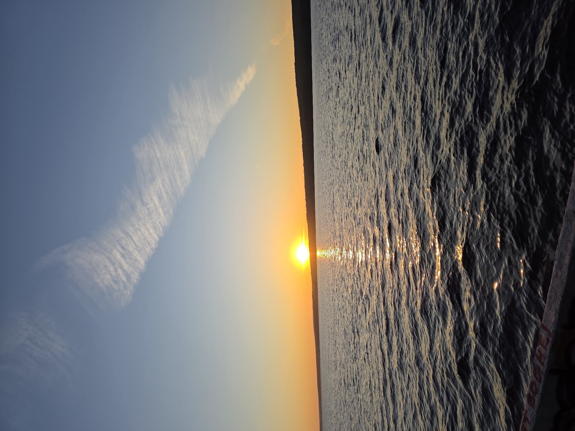 This is a landscape photograph taken at sunset over the Kama River in Chaikovsky, Russia. The sun is low on the horizon, casting a warm golden glow across the water and sky. The sky is a gradient of pale blue and orange, with a wispy cirrus cloud streaking diagonally across the upper half of the frame. The opposite bank of the river is visible as a dark, silhouetted treeline. The water is choppy, with ripples reflecting the sunlight. In the foreground, a wooden structure, possibly part of a dock or pier, is visible at the bottom edge of the image, showing some red lettering. There are no people or animals present in the scene. The overall mood is serene and peaceful.