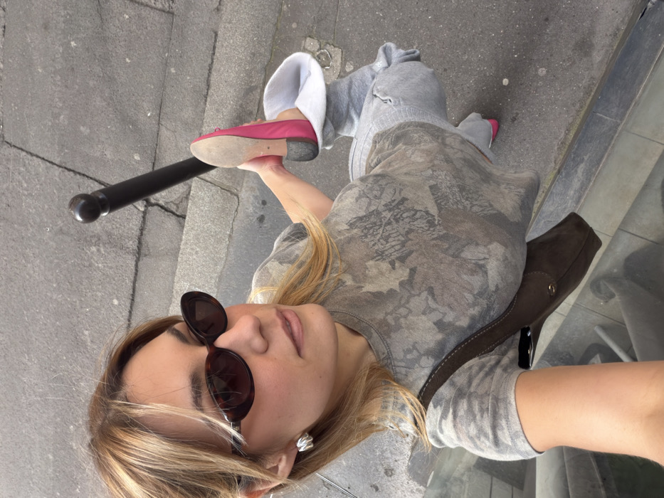 The image displays a high-angle view of a woman posing on a grey asphalt sidewalk in Paris, France. The location is indicated by a black, cylindrical metal bollard with a rounded top, commonly known as a "bornette," against which the woman's raised foot rests.
The subject is a light-skinned woman with medium-length, blonde hair, wearing dark, oval-shaped sunglasses and a silver-toned, abstractly shaped earring on her visible left ear. She is dressed in a short-sleeved t-shirt with a grey and brown camouflage or abstract leaf pattern, paired with light grey sweatpants. Both of her feet are clad in bright pink ballet flats; the sole of her right shoe is visible and appears worn.
The woman is captured mid-pose, with her right leg bent and her right foot lifted to rest against the black bollard. Her left foot is partially visible on the ground. She looks towards the left of the frame with a neutral expression. A dark brown shoulder bag, featuring a metallic buckle and visible stitching, hangs over her left shoulder. The street surface exhibits cracks and variations in texture, and a reflective surface is partially visible in the bottom left corner, possibly a storefront window or vehicle.