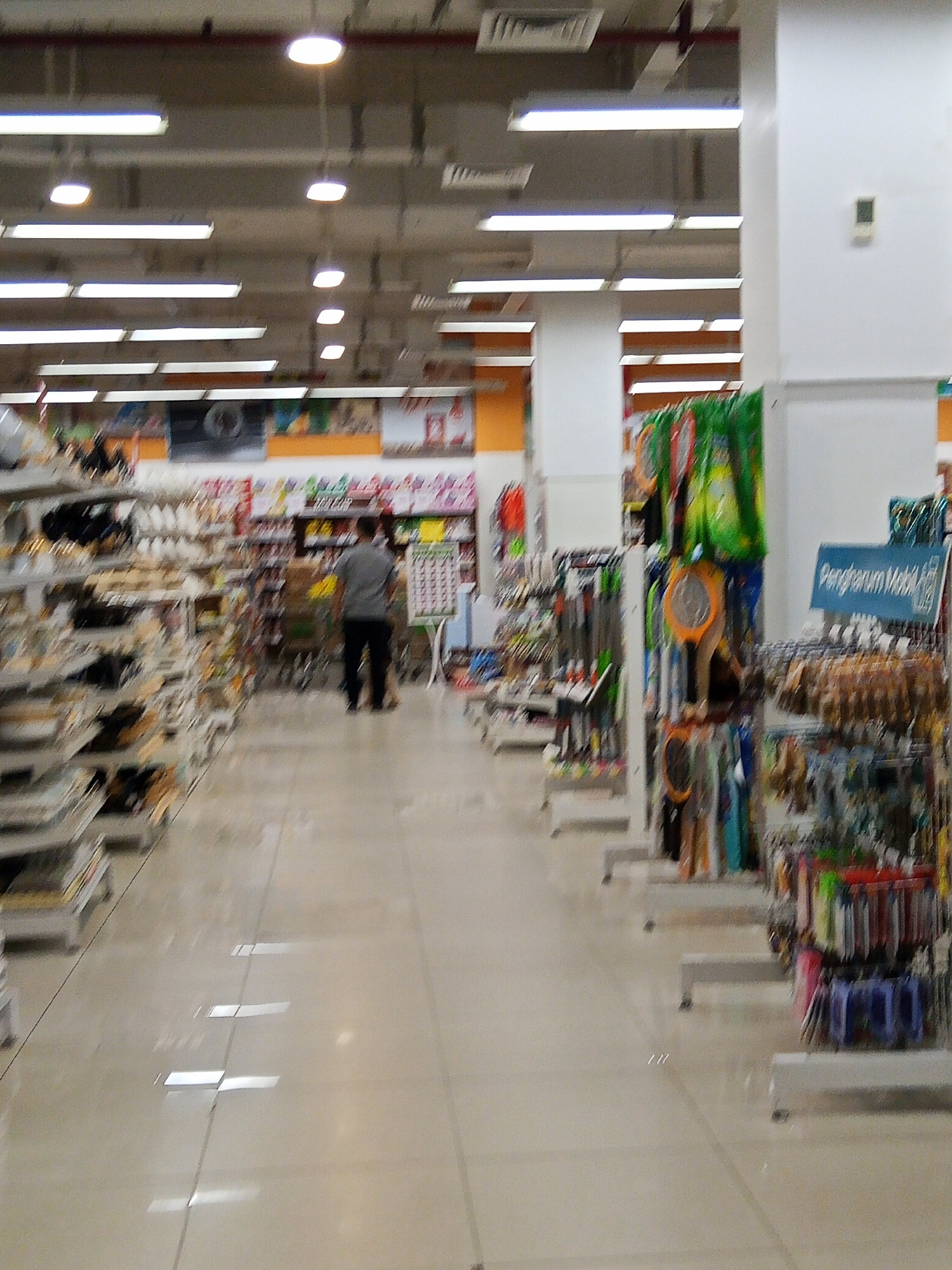 The image shows the interior of a supermarket or department store, likely in Lubuklinggau, Indonesia, given the context of the request. The scene is well-lit by fluorescent lighting from the ceiling. In the foreground, there are aisles with shelves displaying various products, including what appear to be household goods, cleaning supplies, and possibly personal care items. The floor is tiled and reflects the overhead lights.
Further down the aisle, a person is visible walking away from the camera. This individual is wearing dark trousers and a grey t-shirt. They appear to be browsing or walking through the store.
The middle ground and background reveal more aisles, product displays, and shelving units. Some displays feature colorful items like mosquito rackets and car air fresheners, indicated by the "Pengharum Mobil" sign. The overall impression is that of a busy retail environment with a wide array of merchandise. The image is slightly blurred, suggesting it was taken with a moving camera or in low light conditions, contributing to a sense of motion within the scene.