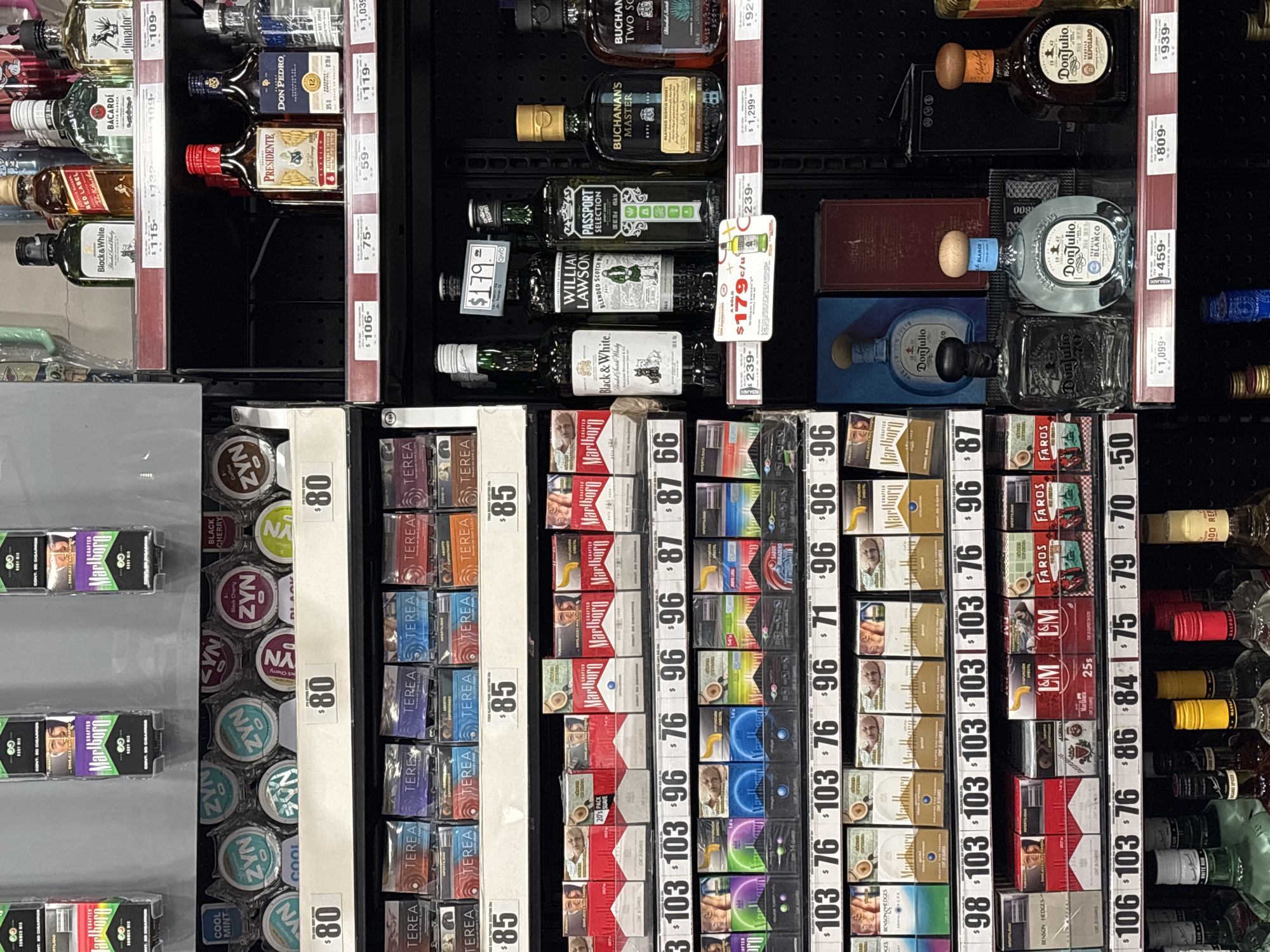 This image captures a well-stocked retail display featuring a variety of alcoholic beverages, cigarettes, and nicotine pouches within what appears to be a convenience store or supermarket aisle. The products are neatly arranged on multiple shelves, with visible price tags for nearly every item.
The top sections of the shelves are dominated by liquor bottles, including brands like El Jimador, Bacardi, Red Label, Presidente, Don Pedro, Buchanan's Two Souls, Buchanan's Master, Passport Selection, William Lawson, Black & White whiskey, and Don Julio (Reposado and Blanco variants). Prices for these bottles range from $59 to $1,299.
Below the liquor, a wide array of tobacco products and nicotine pouches are displayed. Cigarette brands include Marlboro, Faros, L&M, Benson & Hedges, and Markhams, with prices mostly between $50 and $103. Nicotine pouches from ZYN (flavors like Black Cherry and Cool Mint) and Terea are also visible, priced at $80 and $85 respectively. Some price tags indicate "REBAJADO" (reduced) or "c/u" (each one), suggesting sales or individual unit pricing.
The prices, combined with brands common in Latin American markets and the provided context of Puebla, Mexico, indicate that the currency displayed is the Mexican Peso. The scene is static, devoid of people, focusing solely on the products available for purchase.