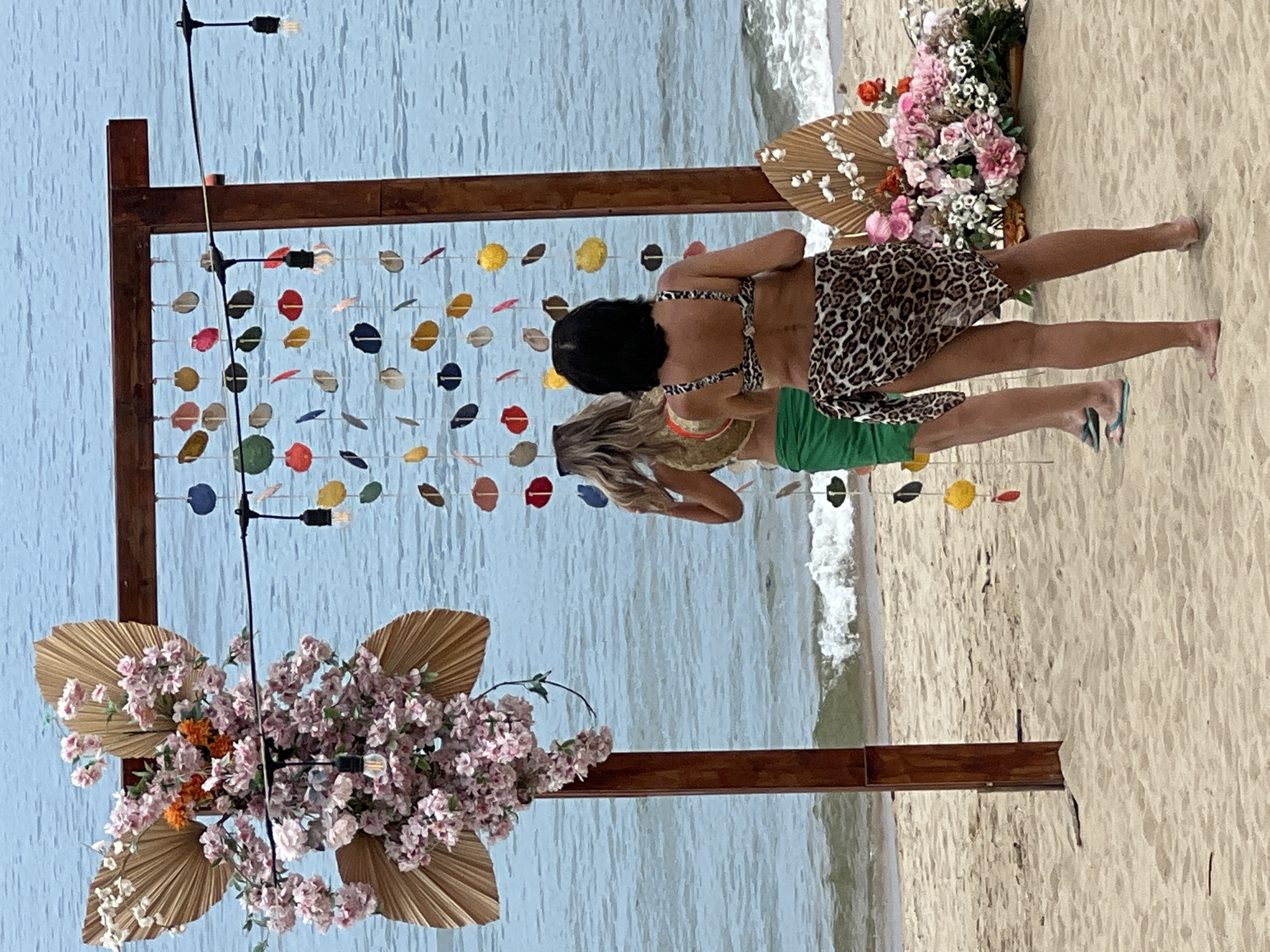 The image depicts a beach scene in Nha Trang, Vietnam, with a wedding arch decorated with pink flowers and dried palm leaves. Two women, dressed in swimwear, stand facing away from the camera, looking out at the ocean. The woman in the foreground wears a leopard print sarong over a bikini, while the woman behind her wears a green sarong over her bikini. A string of colorful shell decorations hangs from the arch, and string lights with exposed bulbs are also visible. The sand is light brown, and gentle waves are breaking on the shore. The overall atmosphere suggests a relaxed, tropical setting.