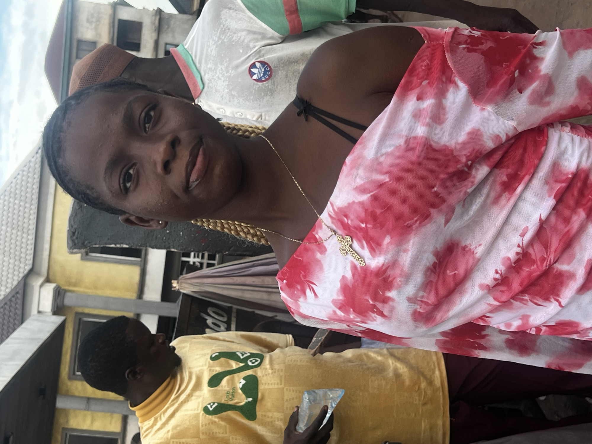 In Nkpor, Nigeria, a close-up shot captures three individuals outdoors. The central subject, a young Black woman, faces the camera, her expression neutral. She is adorned with a gold cross necklace and long braided hair. Her attire consists of a light pink and white tie-dye mesh top with a black bra strap visible. To her left, a Black man in a bright yellow long-sleeved shirt with a green and white graphic logo holds a clear plastic bottle of water. Behind him, a Black man wearing a light-colored polo shirt with green and red trim and a brown patterned cap is partially visible. The background features a building with yellow and grey siding and tiled roofs, suggesting a typical urban environment. The lighting is natural, indicating an outdoor setting during daylight hours. The composition focuses on the central subject, with the other individuals and the environment serving as contextual elements.