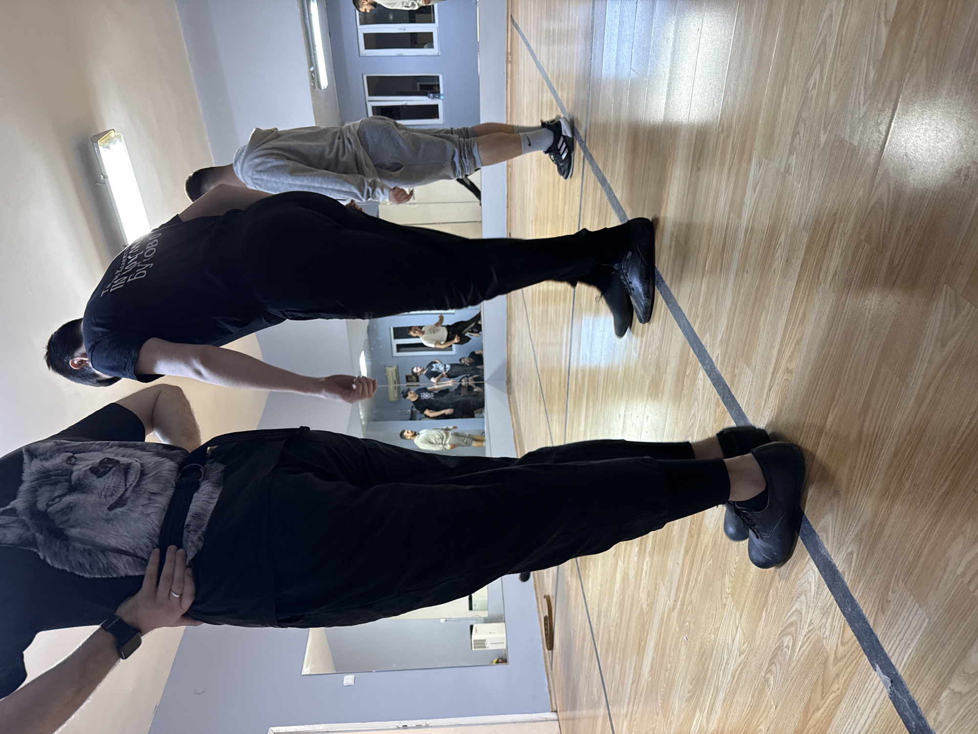 The image displays an indoor scene, likely a training studio or gym, characterized by a light wooden floor with a dark line marking, light-colored walls, and large mirrors reflecting parts of the room and people. Fluorescent lights are visible on the ceiling.
Three individuals are prominent in the foreground, standing upright. The person closest to the camera is wearing a black t-shirt with a detailed wolf graphic, black pants, black shoes, a watch on their left wrist, and a ring on their left ring finger. Their hands are placed on their hips. Behind them, another individual wears a black t-shirt with white Cyrillic text, black pants, and black shoes. The text on the t-shirt reads "ПОБЕДА ДУХА РУКОВОДИТ БОЕМ," which translates to "Victory of the Spirit Guides the Battle." Further back, a third person is dressed in a grey hoodie, grey shorts, and black athletic shoes with socks.
Reflections in the mirrors reveal additional people in the background, suggesting a group activity or class in progress. The individuals appear to be standing in a focused manner, possibly awaiting instructions or holding a pose during a physical training session. The setting, attire, and presence of mirrors are consistent with a dance, martial arts, or fitness class. The artificial lighting indicates an indoor environment. The location is Buhovo, Bulgaria.