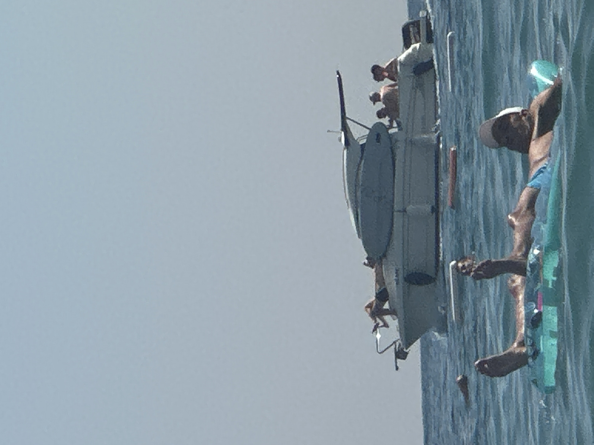 In Boreti, Montenegro, a clear, bright day is captured with a boat and people enjoying the water. In the foreground, a person lies on a teal inflatable ring, their legs and torso visible as they relax on the calm sea. Their feet are pointed upwards, and they wear blue swim trunks and a baseball cap. Behind them, a white motorboat is anchored. Several individuals are on the boat, some appearing to be sunbathing on the deck and others seated in the cabin. The water is a deep blue, with slight ripples indicating a gentle breeze. The sky is a pale, hazy blue, suggesting warm weather.