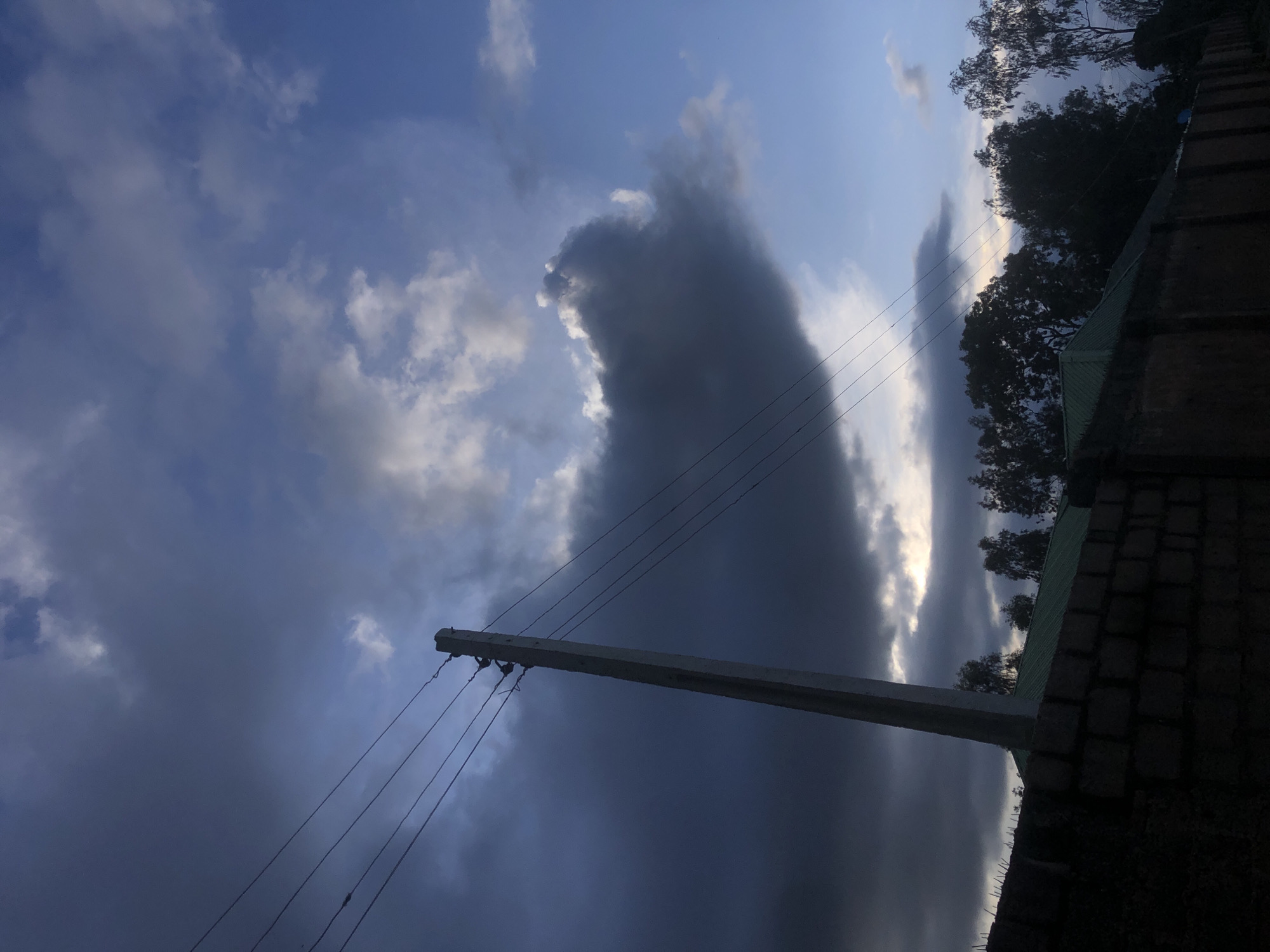 The image shows a scene in Barakin Lerawi, Nigeria, dominated by a dramatic sky with dark, heavy clouds contrasting with patches of blue sky. A concrete utility pole, angled from the lower left towards the upper center, supports multiple overhead wires that stretch horizontally across the frame. Below the pole and wires, a brick wall with a rough texture forms the foreground, with sharp spikes or points visible along its top edge. Beyond the wall, the silhouettes of trees and buildings with green roofs are visible against the sky, with the overall lighting suggesting either early morning or late evening.
