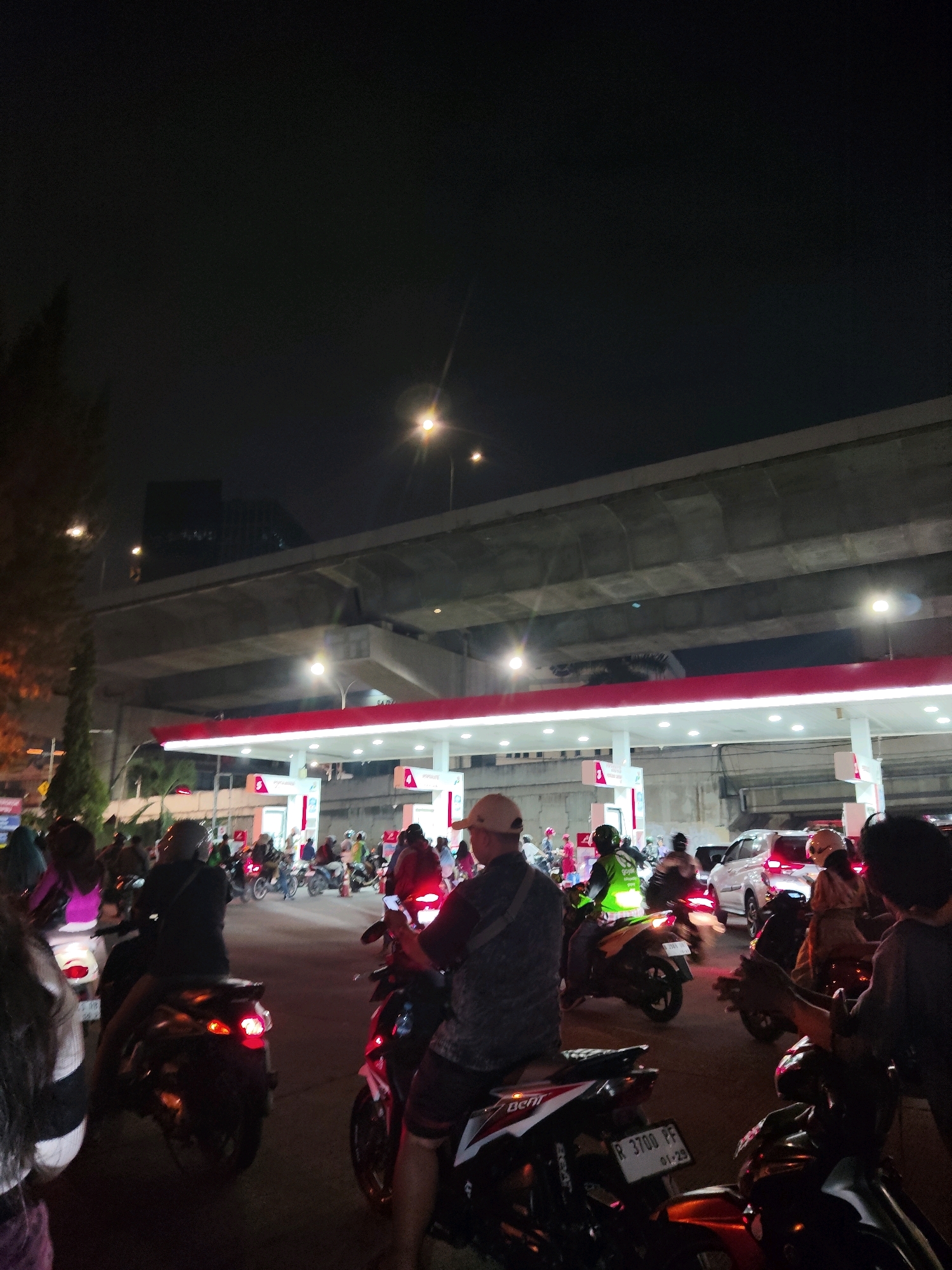 This is a low-light, vertical, first-person perspective image taken at night in North Jakarta, Indonesia. The scene depicts a busy street scene at a gas station, likely part of a Pertamina station given the red and white branding and logo visible. Numerous motorcycles and a few cars are present, suggesting traffic congestion or a gathering. Several people are on or near motorcycles, some wearing helmets and some in casual attire. The background features an elevated concrete structure, possibly a highway overpass, with streetlights illuminating the area. The overall atmosphere is urban and active.